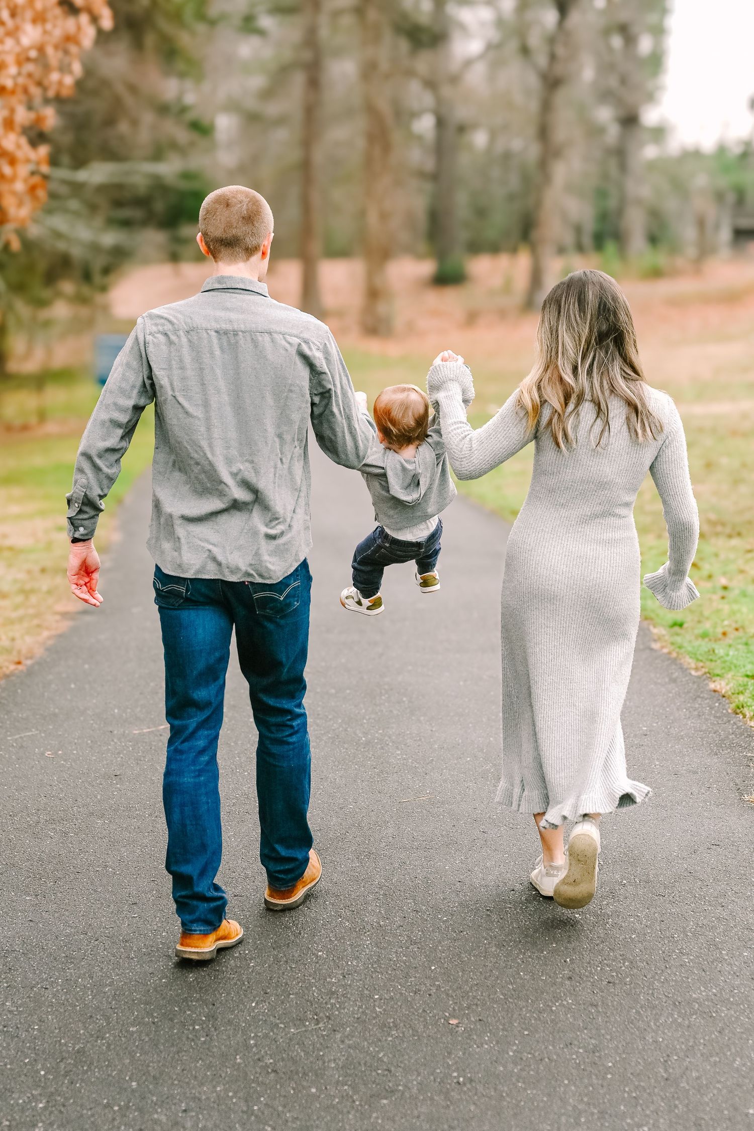 A couple walks down a park path while swinging their small child between them in an autumn setting.
