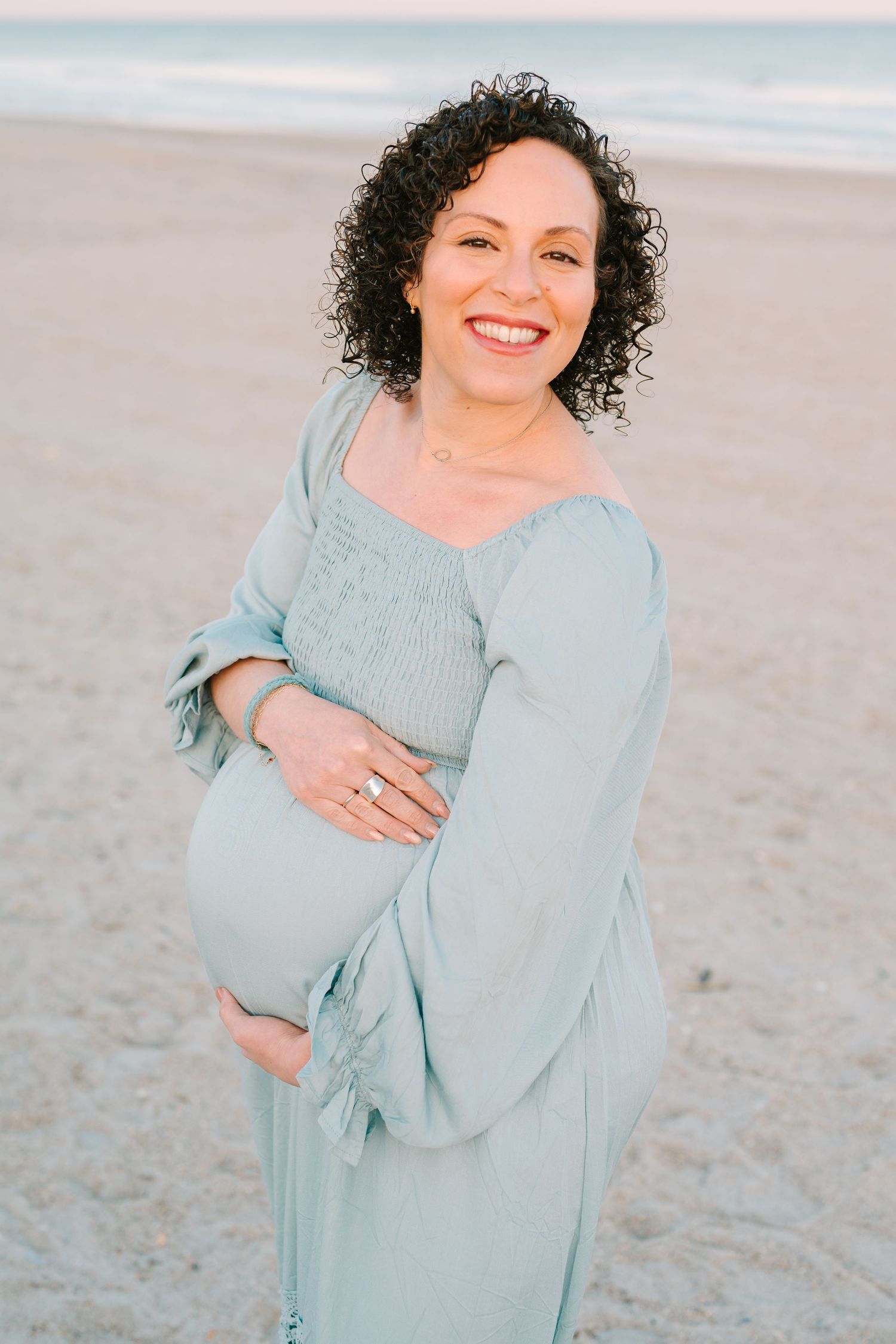 A pregnant woman in a light blue dress stands smiling on a beach at sunset with natural curly hair.