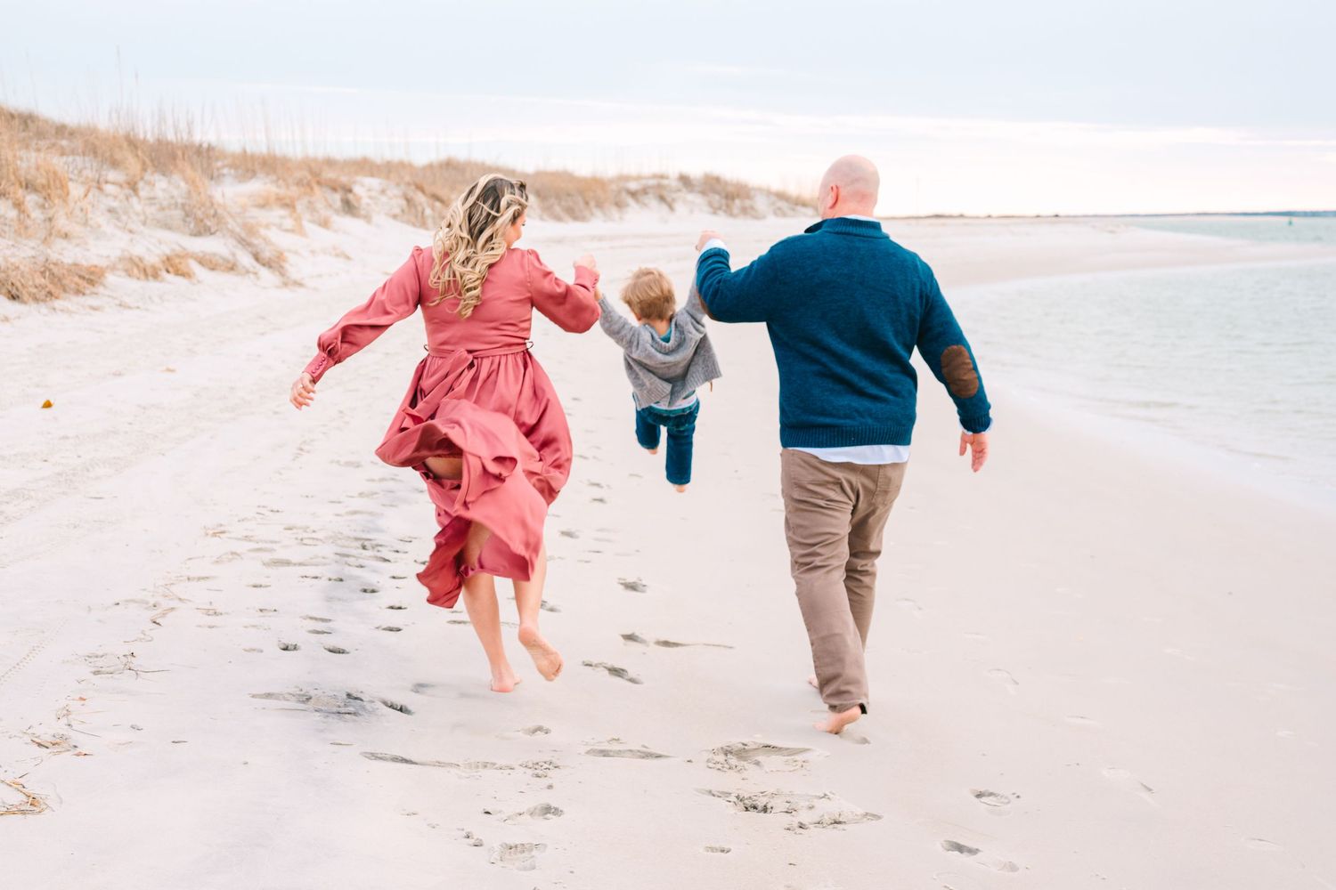 Three people run and play together along a sandy beach on a bright sunny day by the ocean.