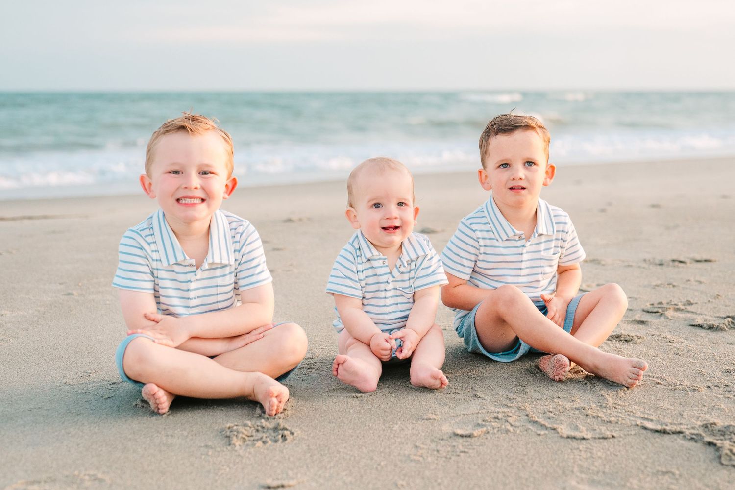 Three young children in matching striped shirts sitting on the sand at the beach.