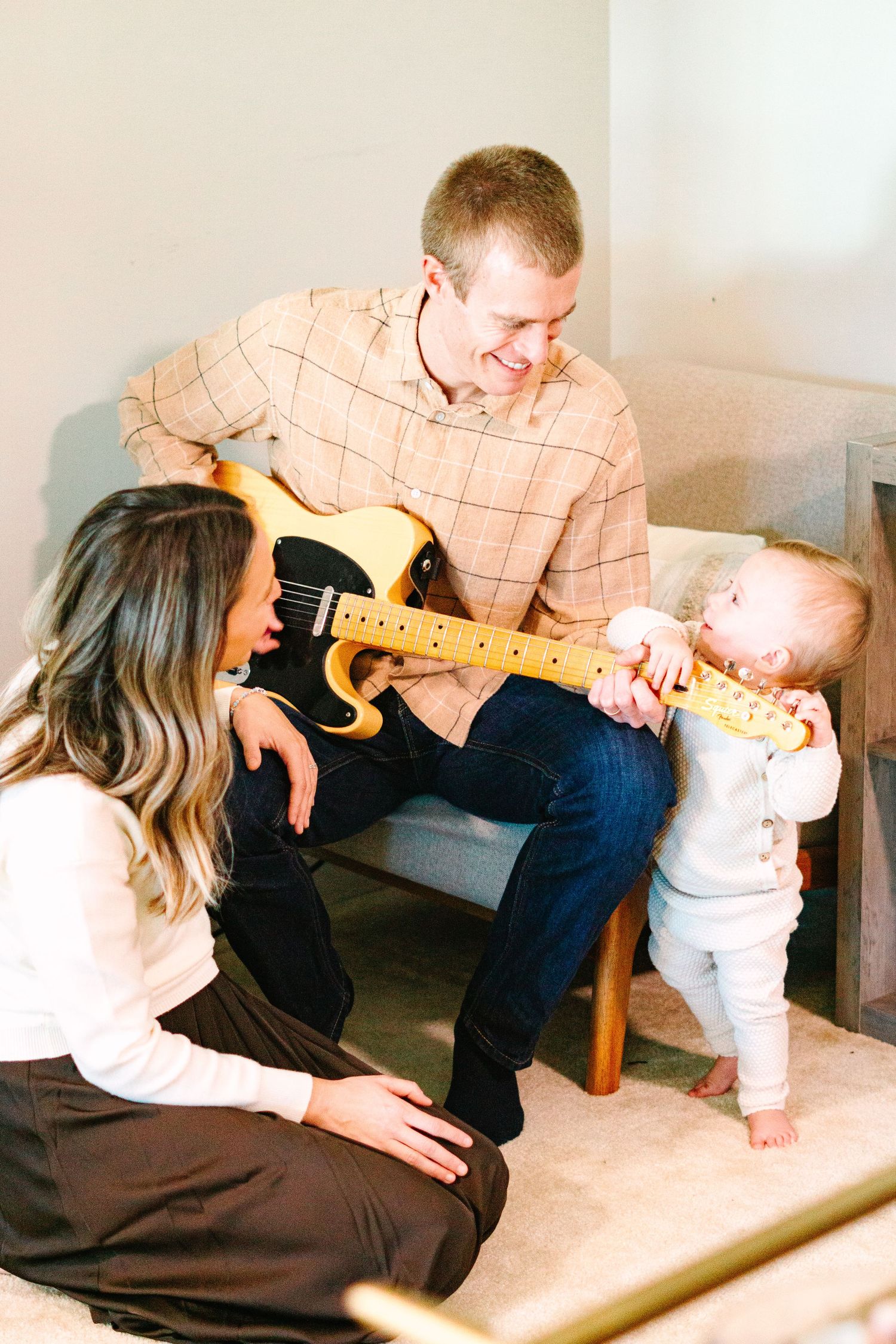 Family plays guitar together in living room as baby reaches for instrument.