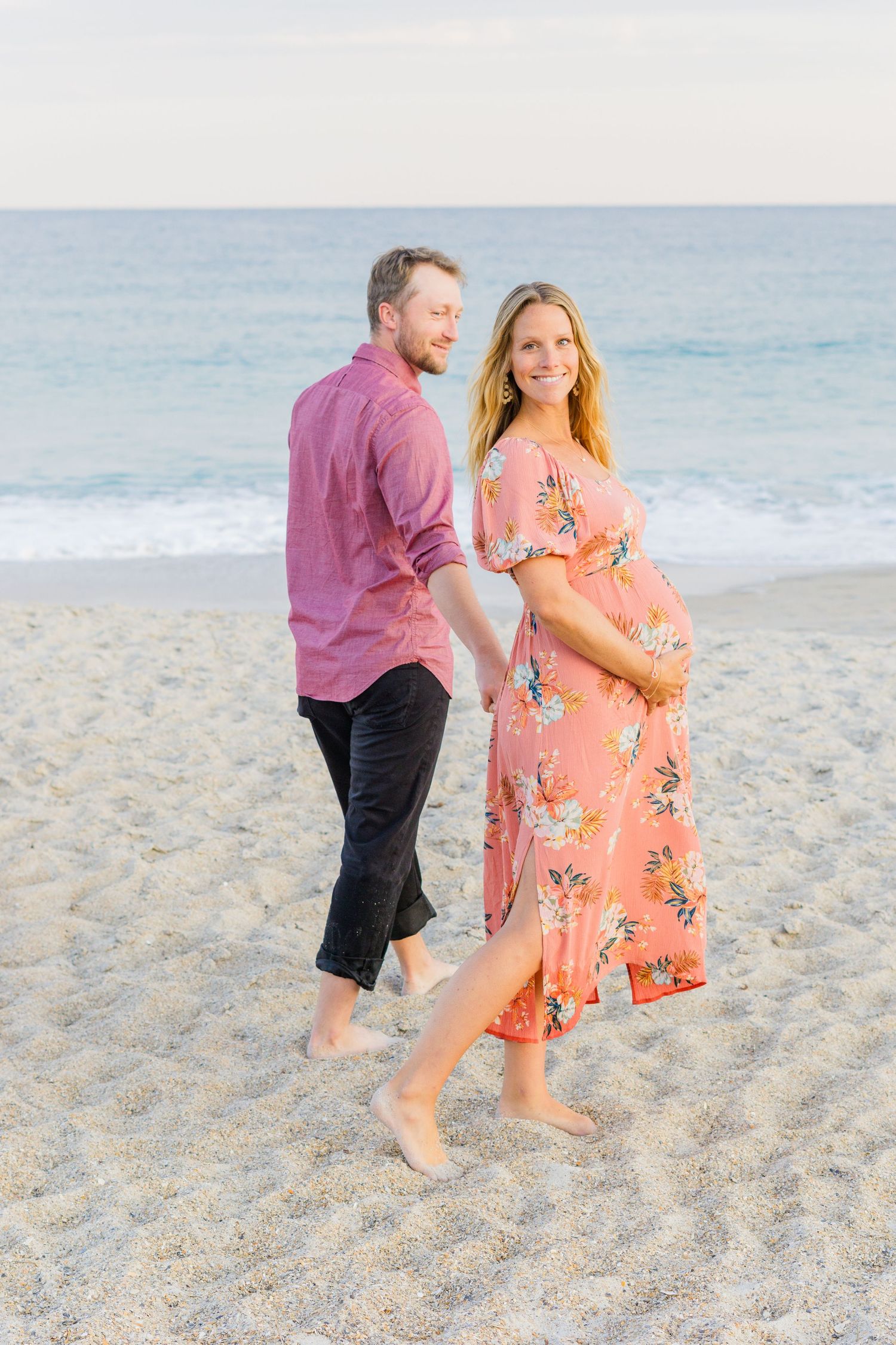 Couple walking barefoot on beach at sunset with woman wearing coral floral maternity dress.