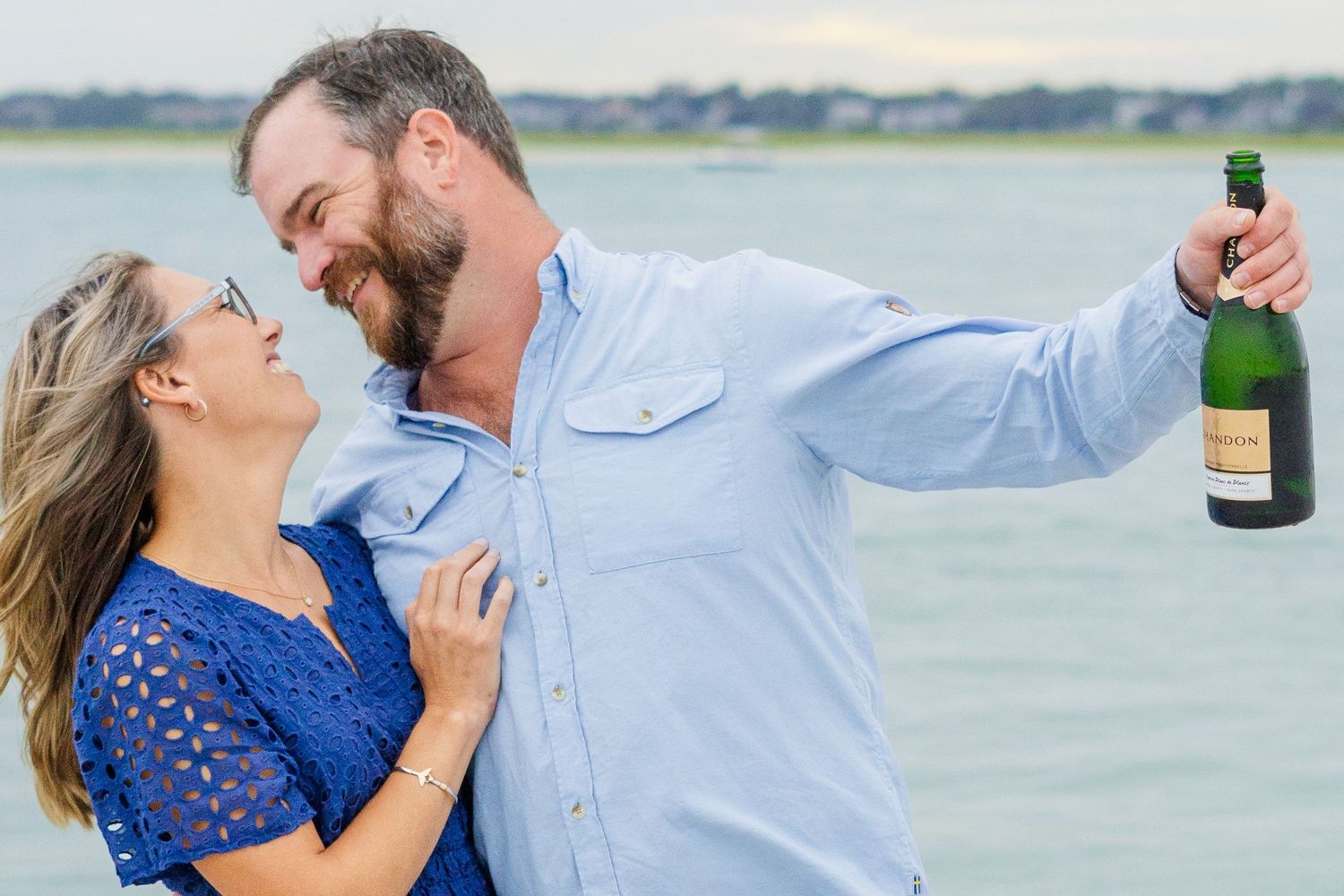 Couple shares a playful moment on the beach while holding a champagne bottle against a waterfront backdrop.