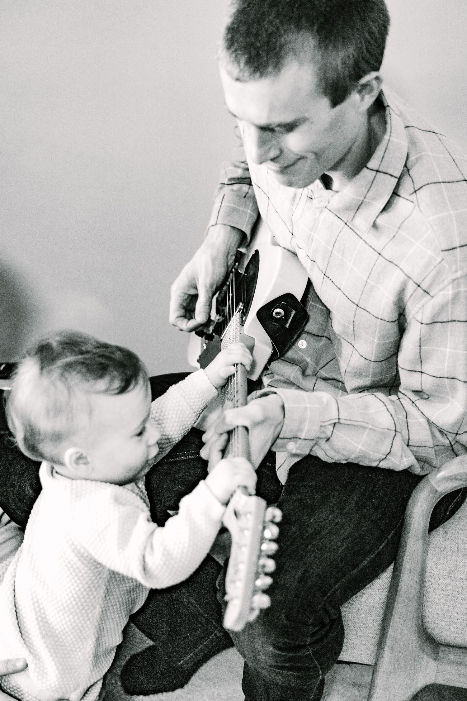 Black and white image of parent and baby exploring guitar strings together.