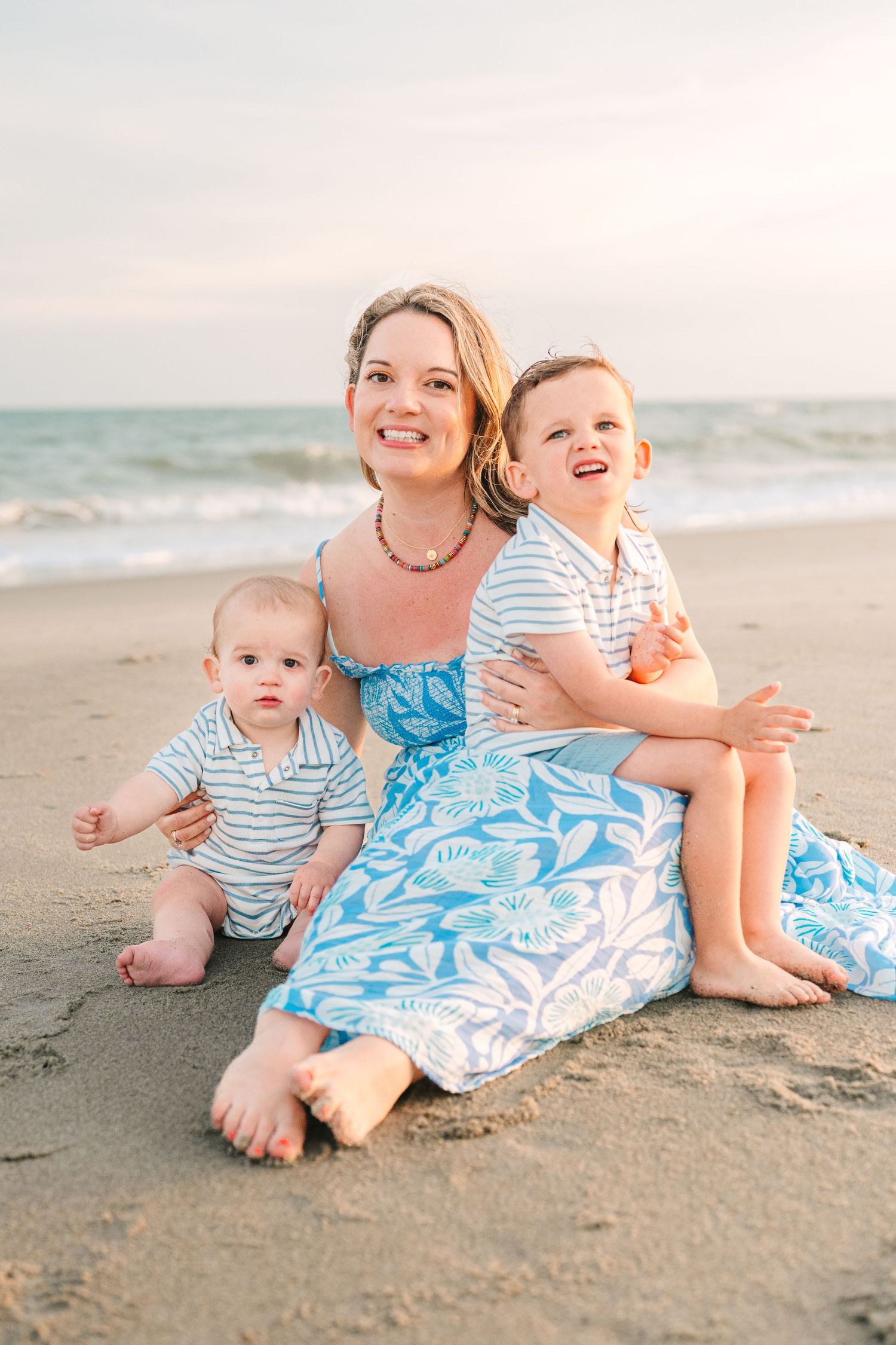 Beach family portrait of mother and children sitting in sand wearing coordinating blue and white outfits.
