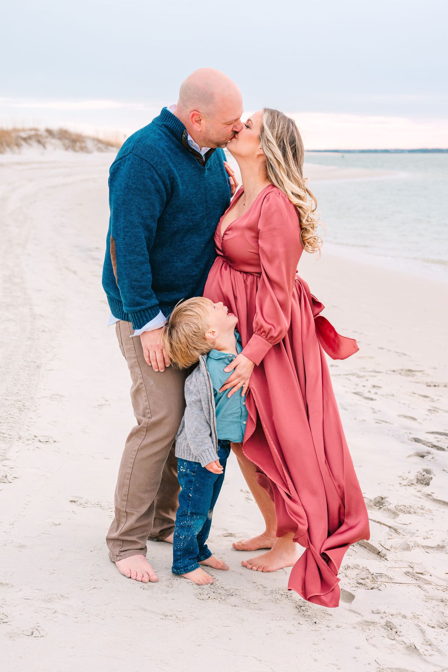A couple shares a romantic moment on the beach while their small child hugs their legs.