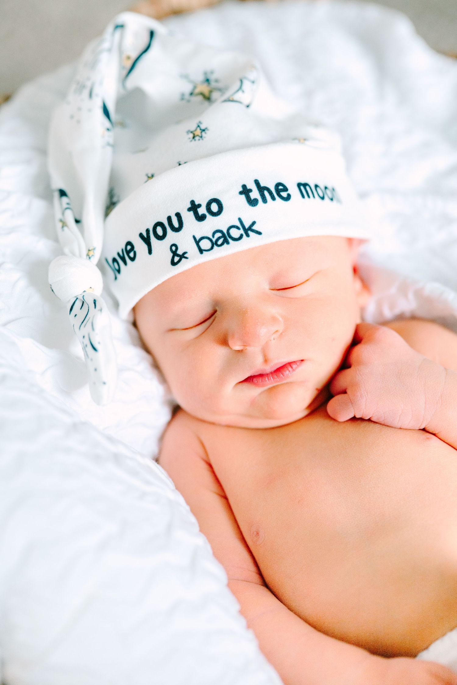 Close-up of peaceful sleeping newborn wearing white hat with moon and stars design on soft bedding.