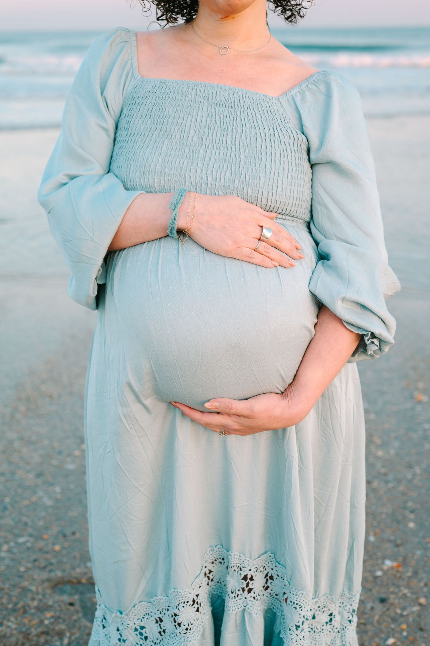 Pregnant woman in light blue dress with smocked bodice and lace trim stands on beach at sunset.