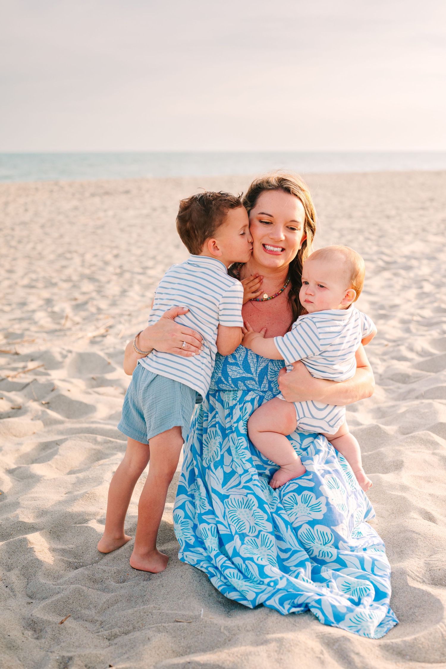 Woman in blue patterned dress embraces two young children on a sunny beach at sunset.