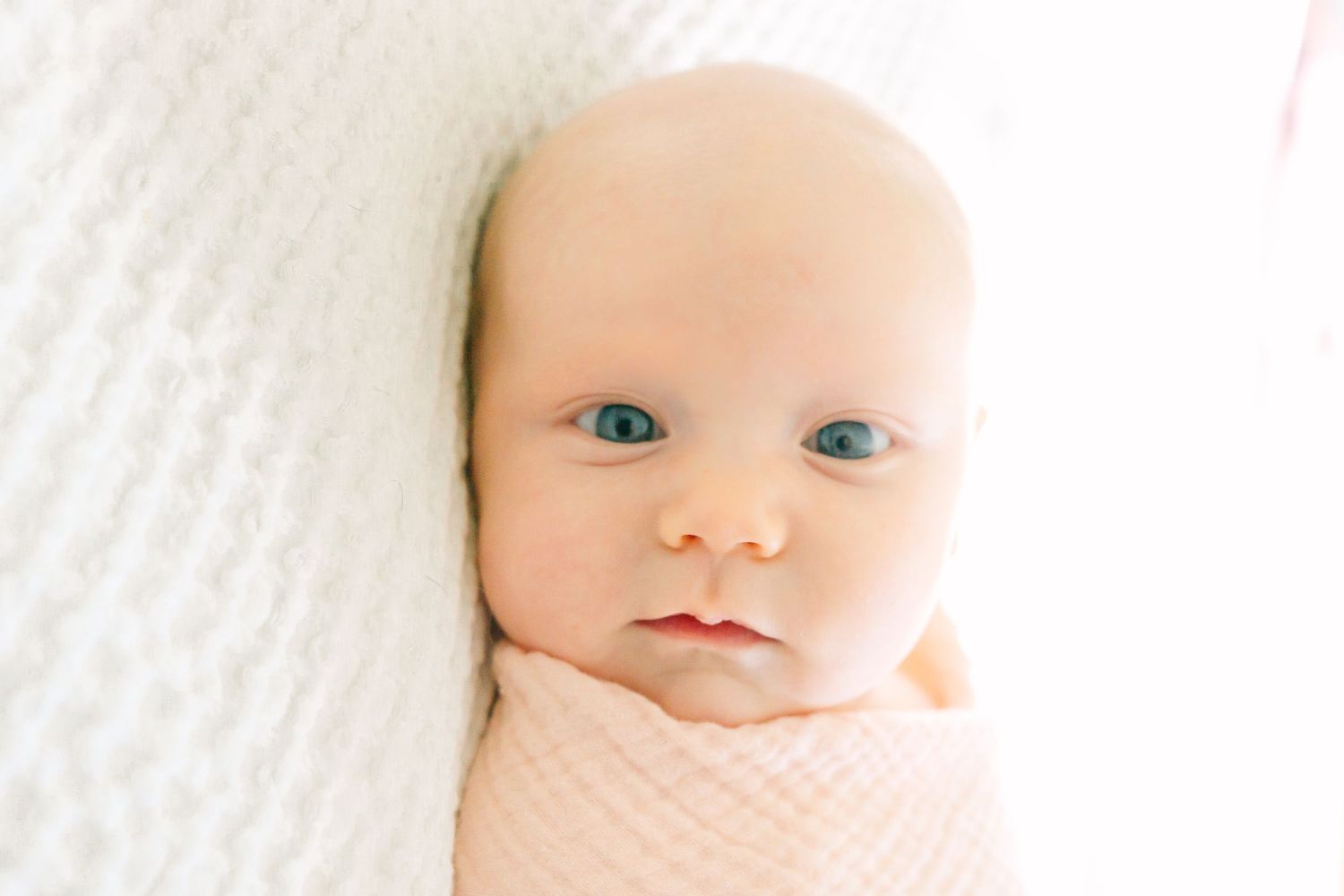Sweet infant swaddled in pink laying on white blanket with bright blue eyes and soft features.