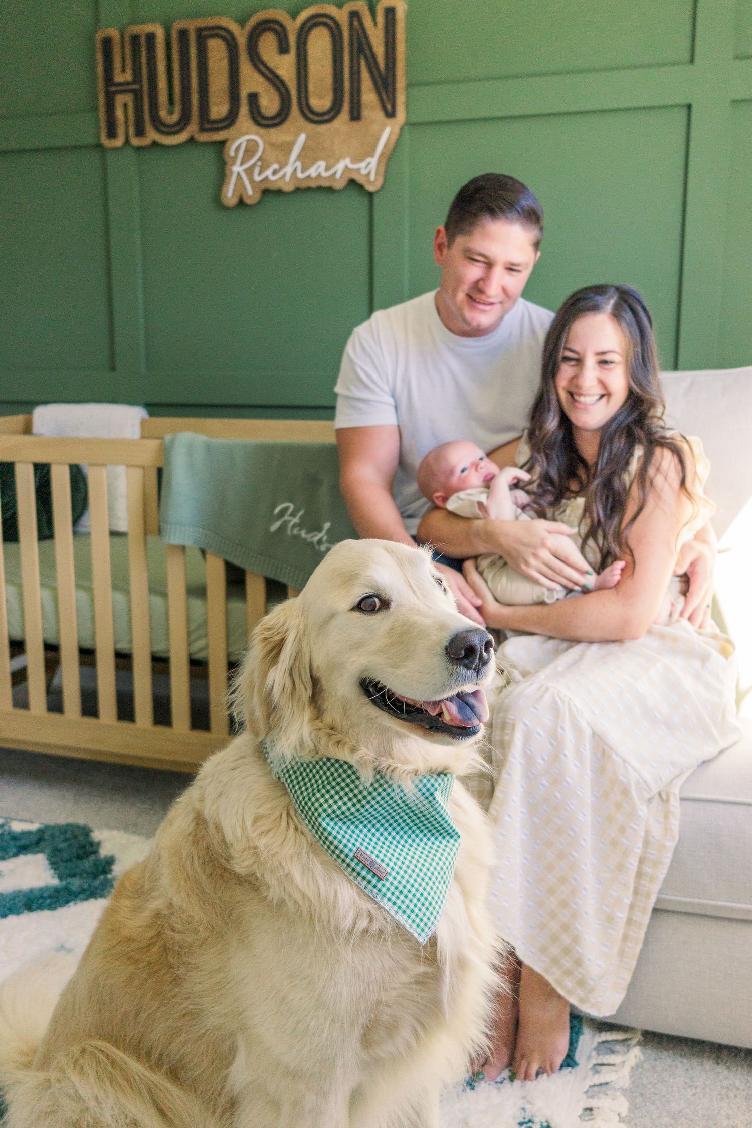 Happy couple with newborn and golden retriever in front of green wall with Hudson sign.