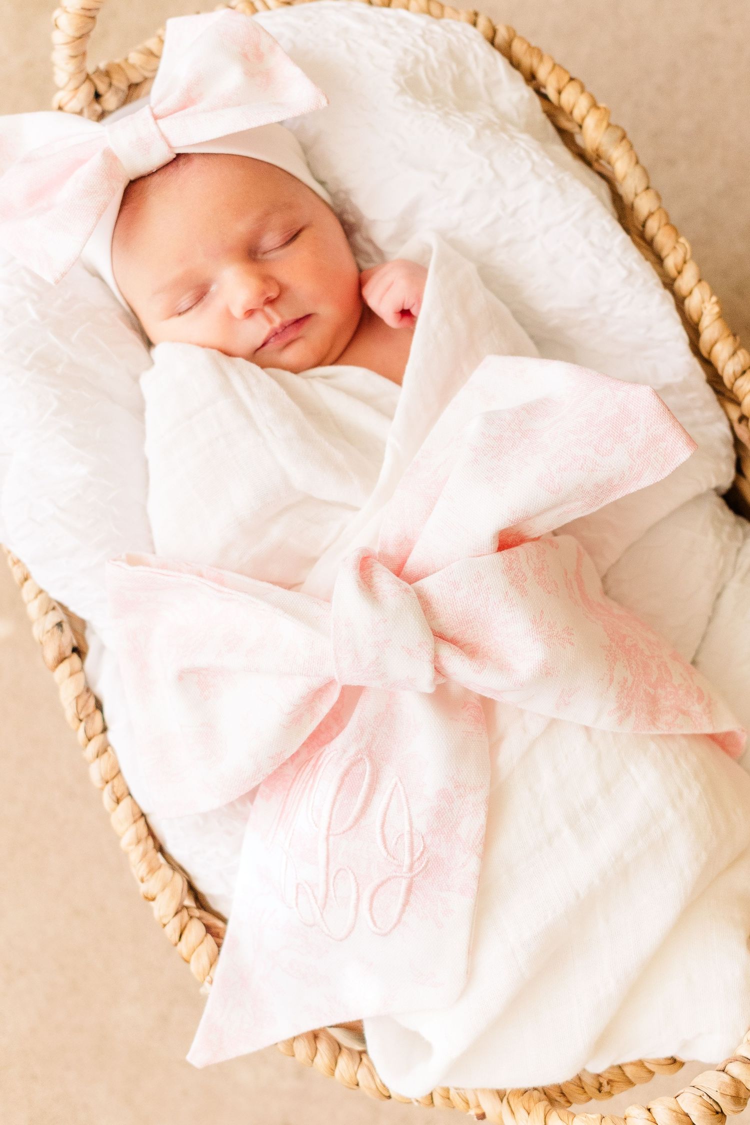 Sleeping newborn wrapped in white blanket with pink bow in woven basket.