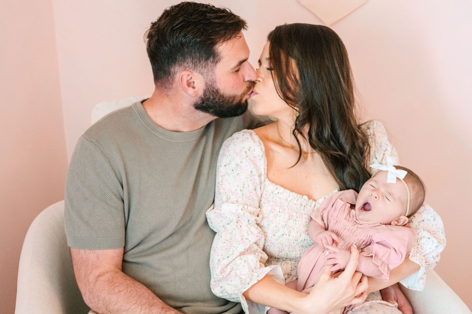 A couple shares an intimate moment while holding their baby in a pink nursery setting.