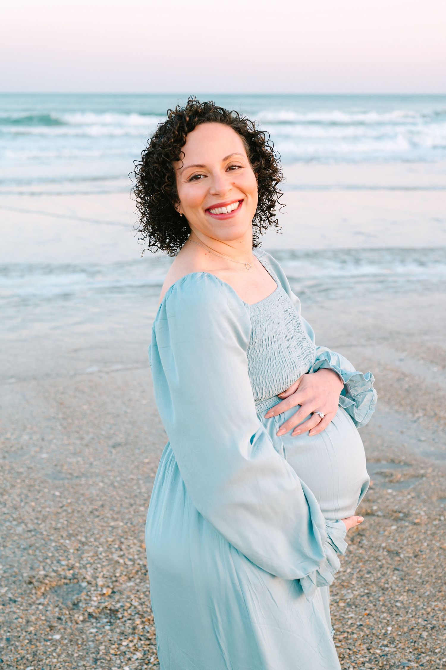 Expecting mother in a light blue dress stands smiling on a beach at sunset with gentle waves in the background.