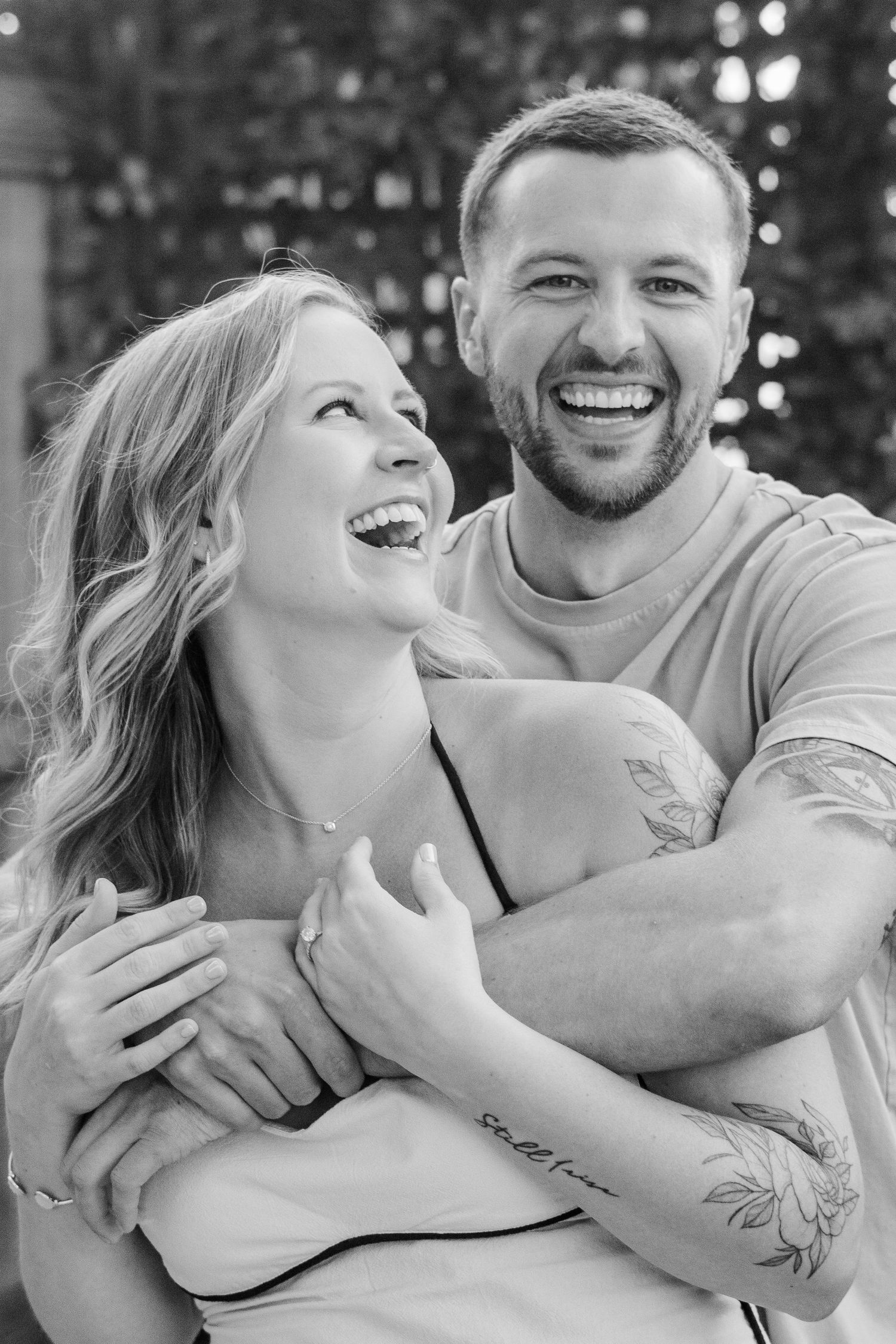 Black and white portrait of couple sharing laughter against natural foliage backdrop.