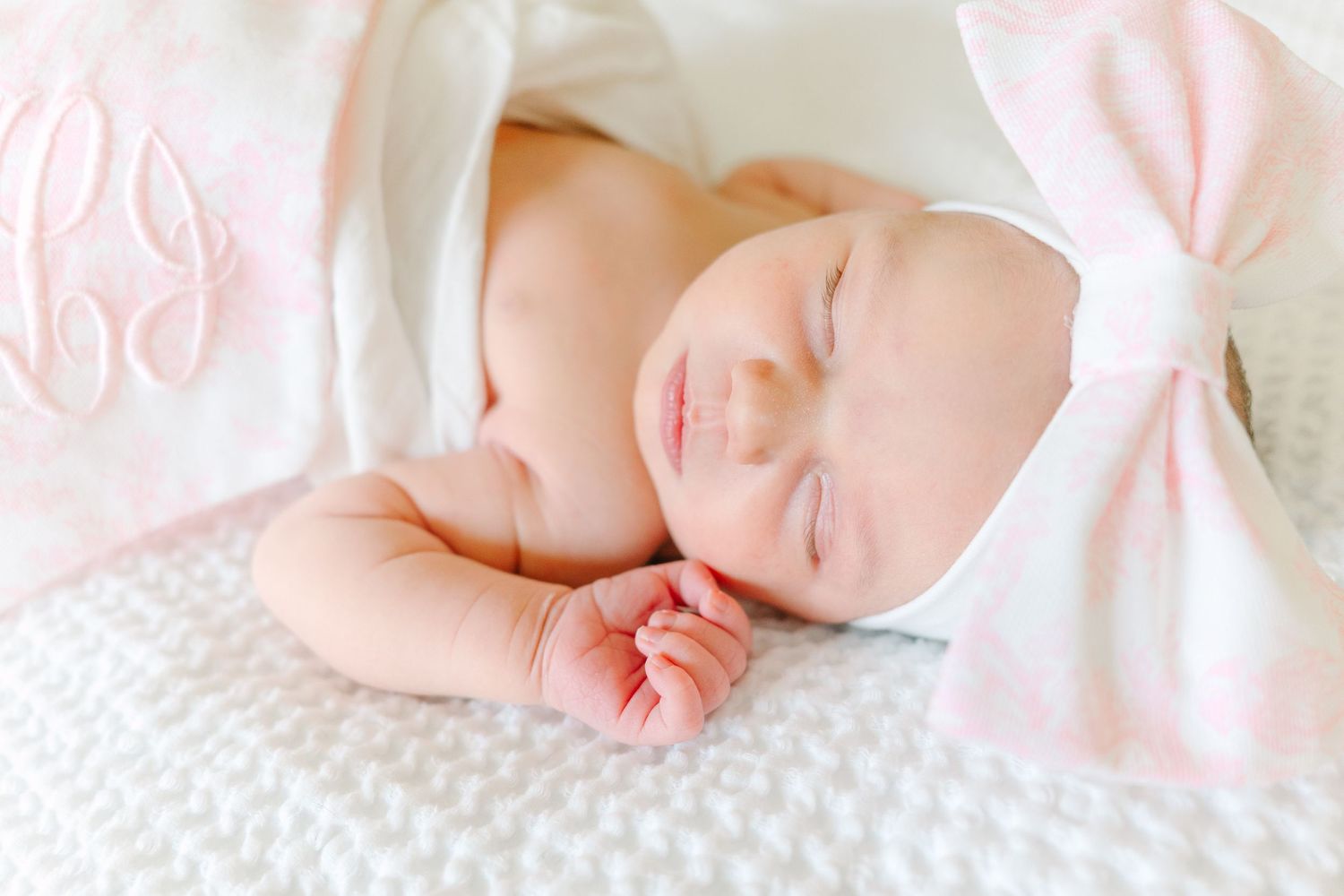 Sleeping newborn wearing white bonnet and wrapped in monogrammed blanket on textured white bedding.