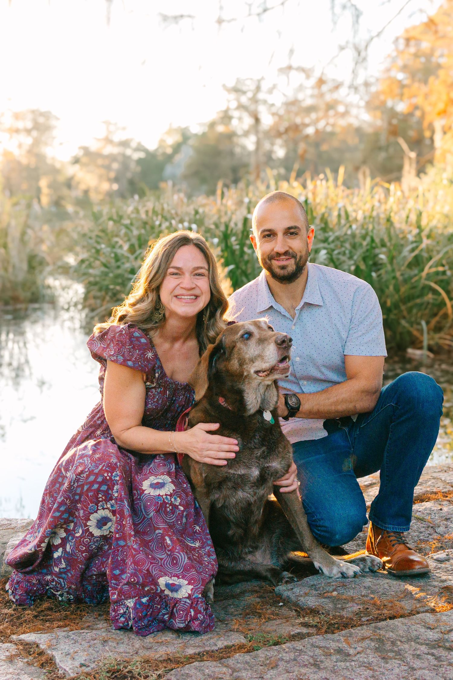 A couple poses with their brown dog by a waterfront during golden hour surrounded by autumn foliage.