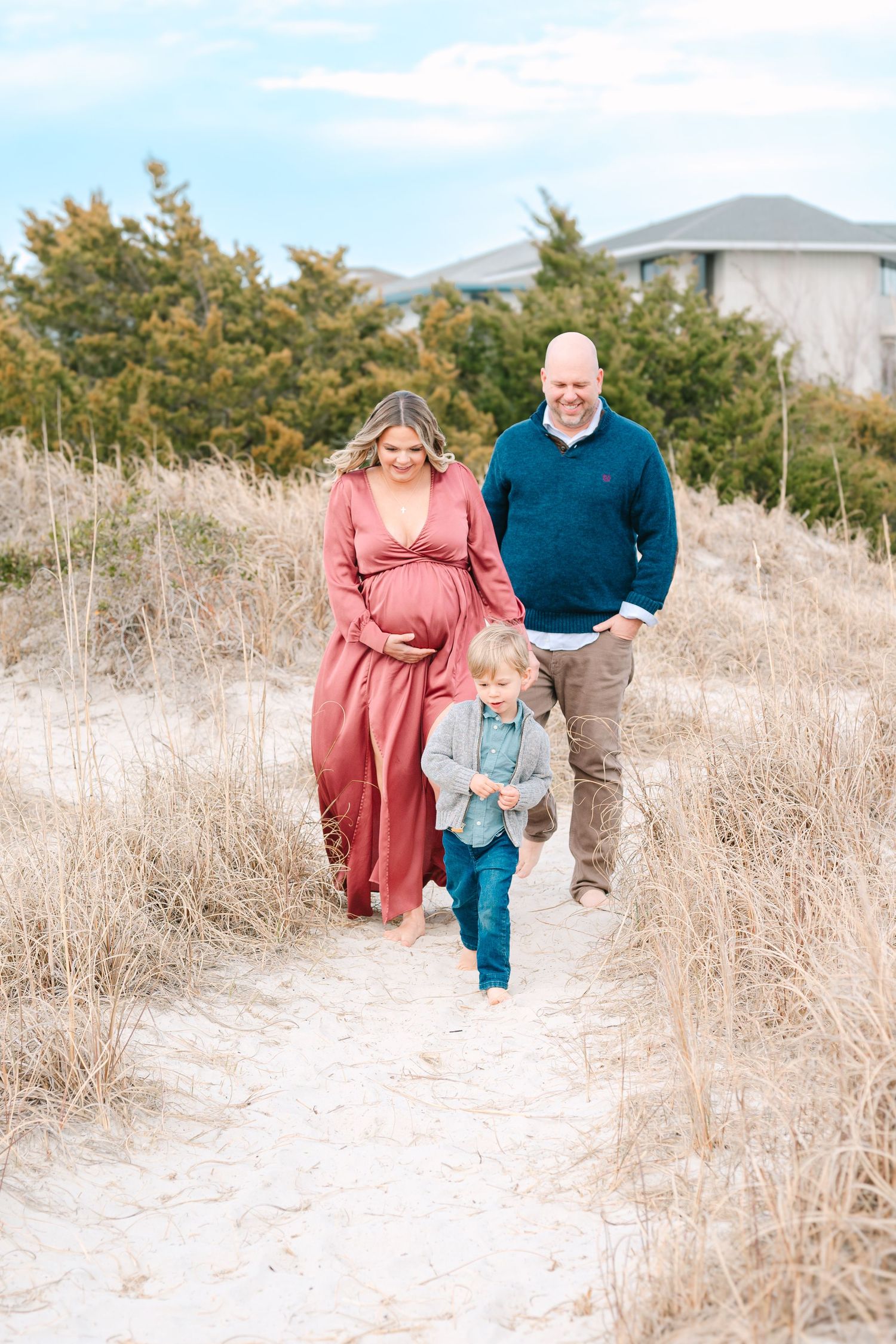 A family walks together on a sandy path through tall grass with evergreen trees and houses in the background.