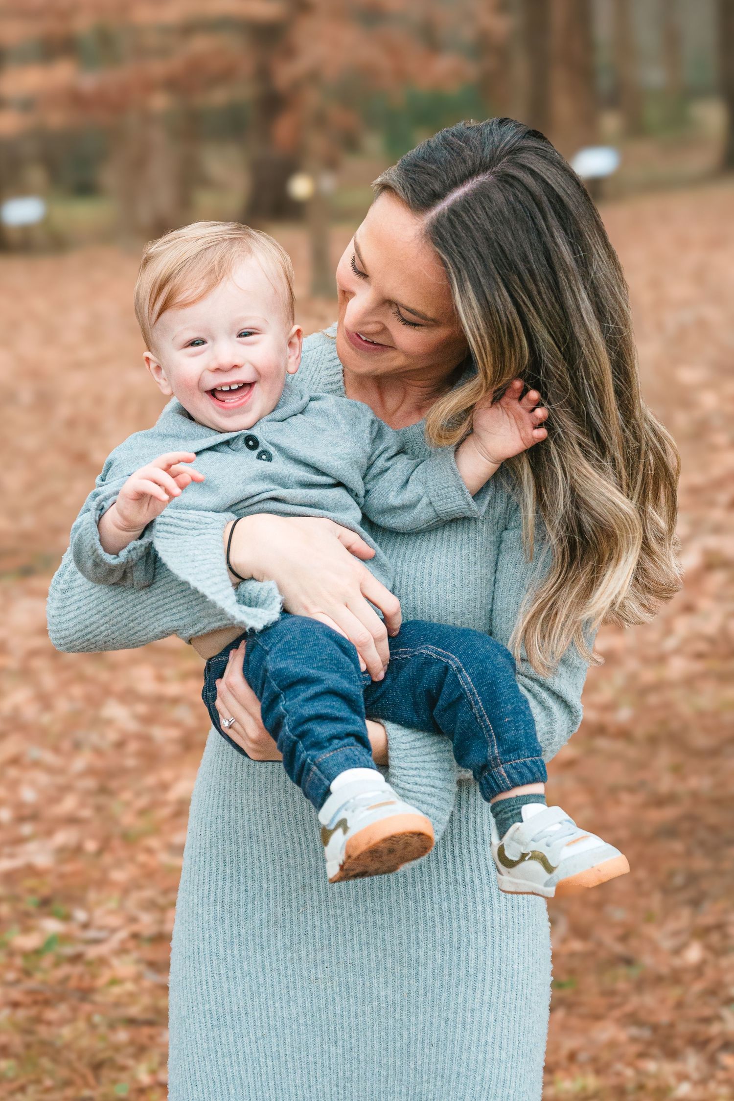 A parent in a gray sweater dress holds and smiles at a laughing toddler wearing blue denim against a backdrop of autumn leaves.