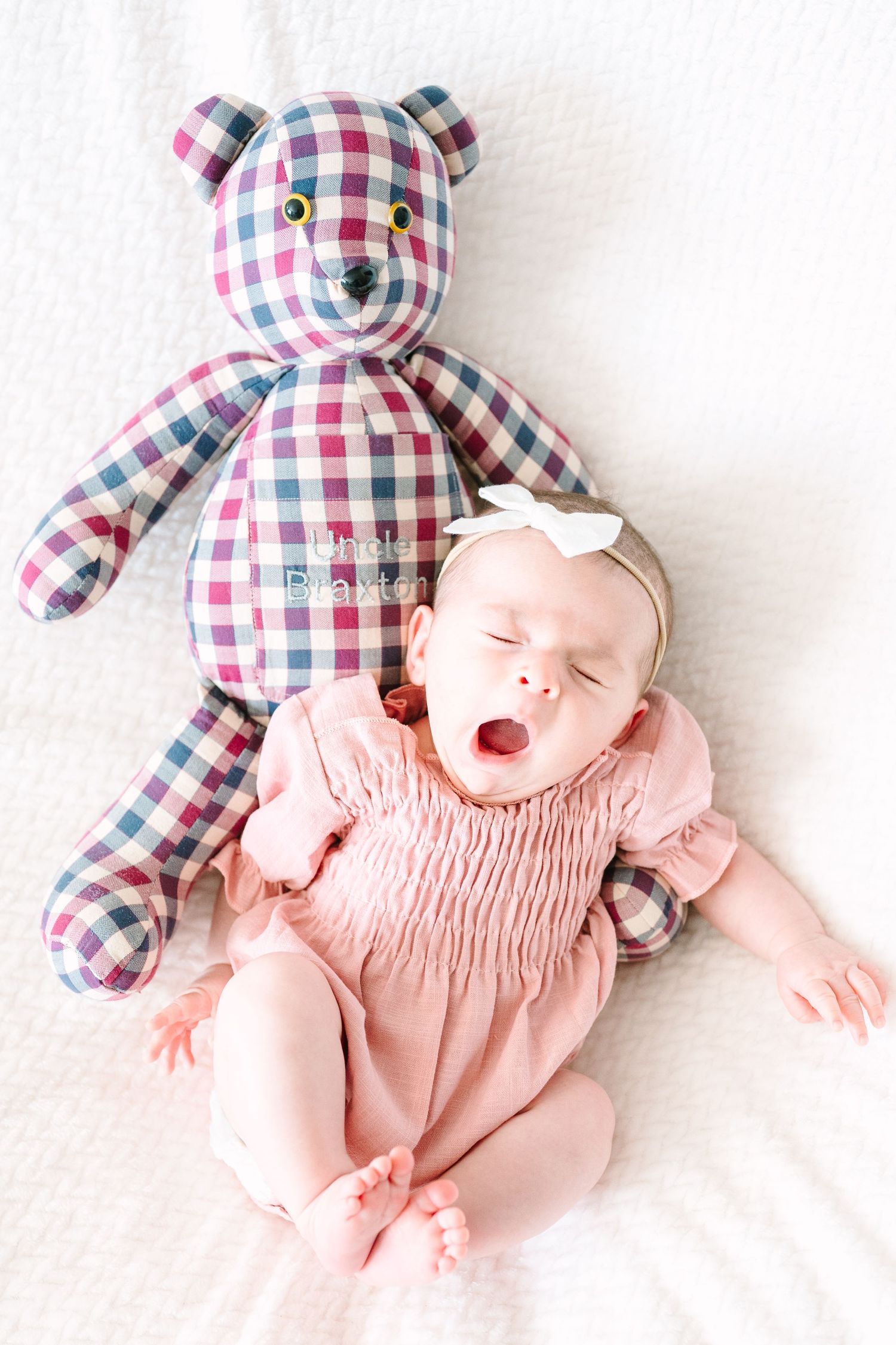 A sleeping baby in pink smocked dress lies next to a large plaid teddy bear on white bedding.