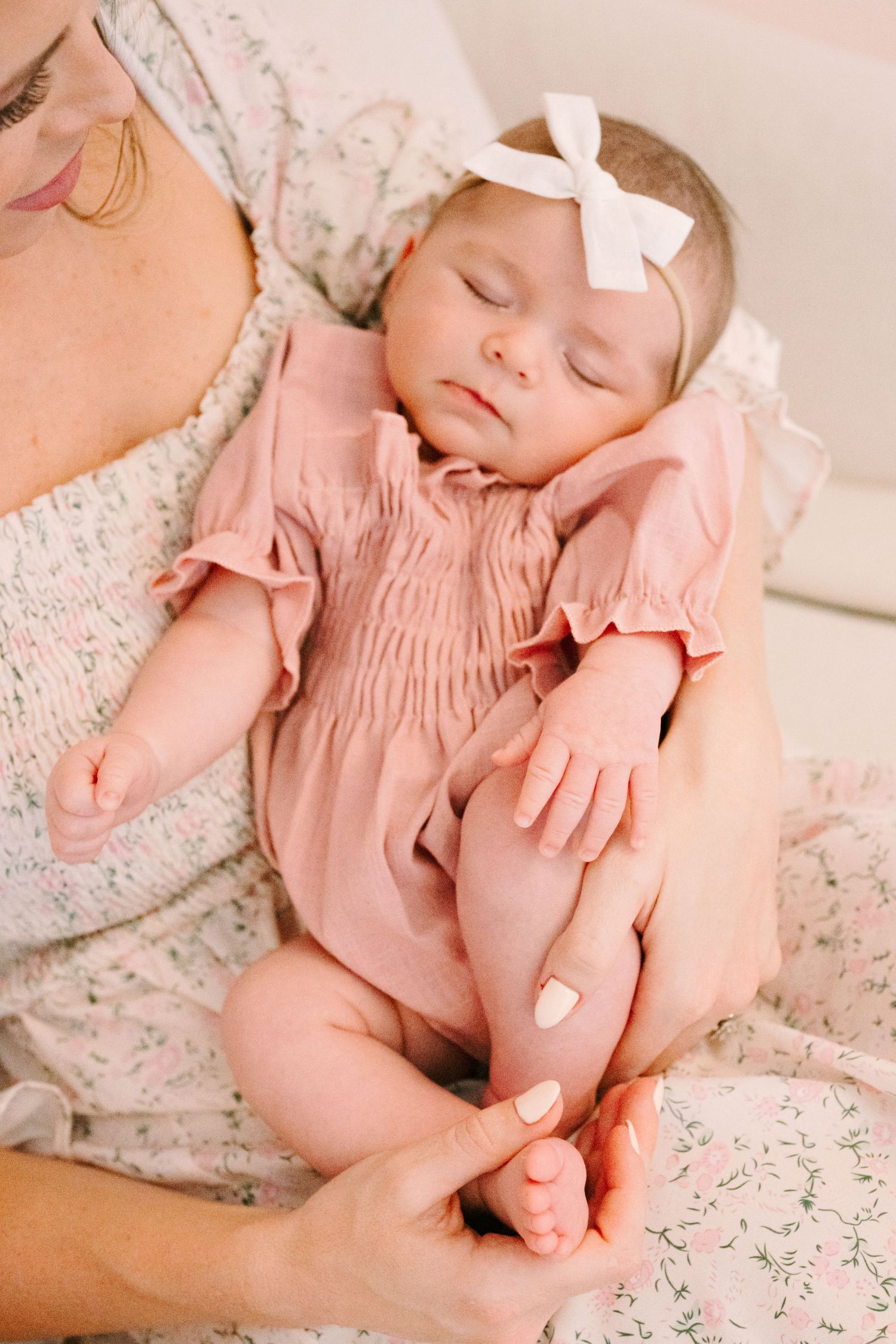 Sleeping newborn baby with white bow headband wearing pink smocked outfit resting on caregiver's chest.