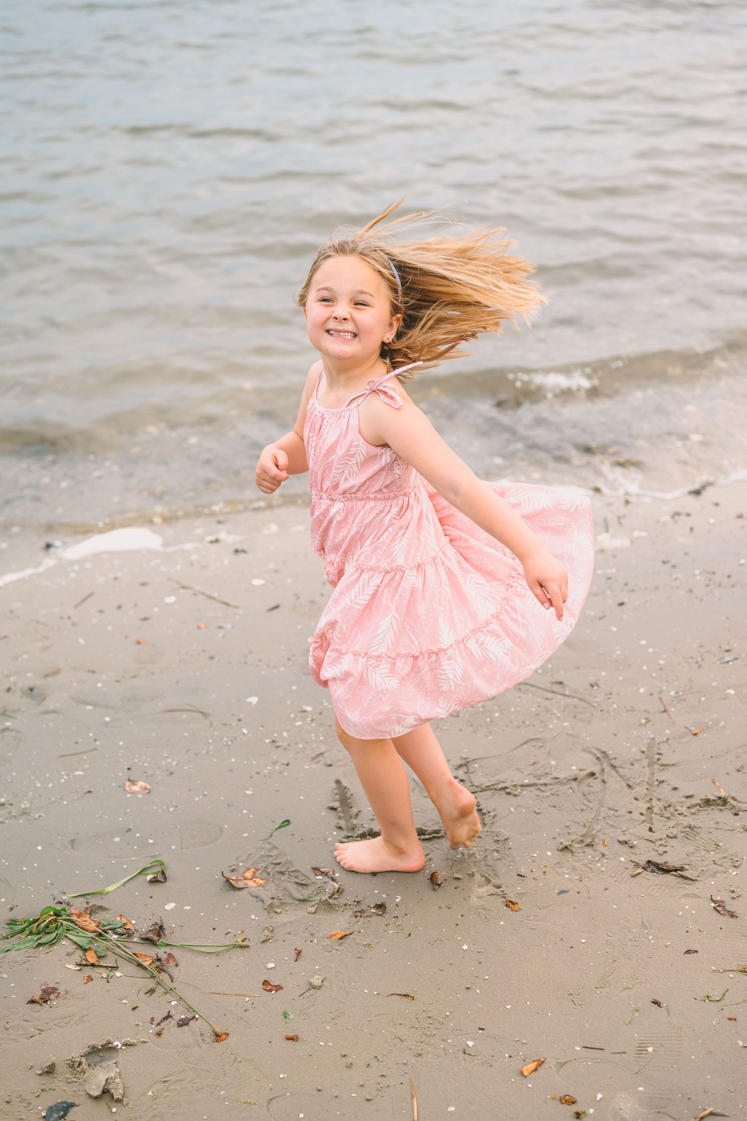 A carefree child twirls in a pink dress on a beach with their hair flowing in the wind.