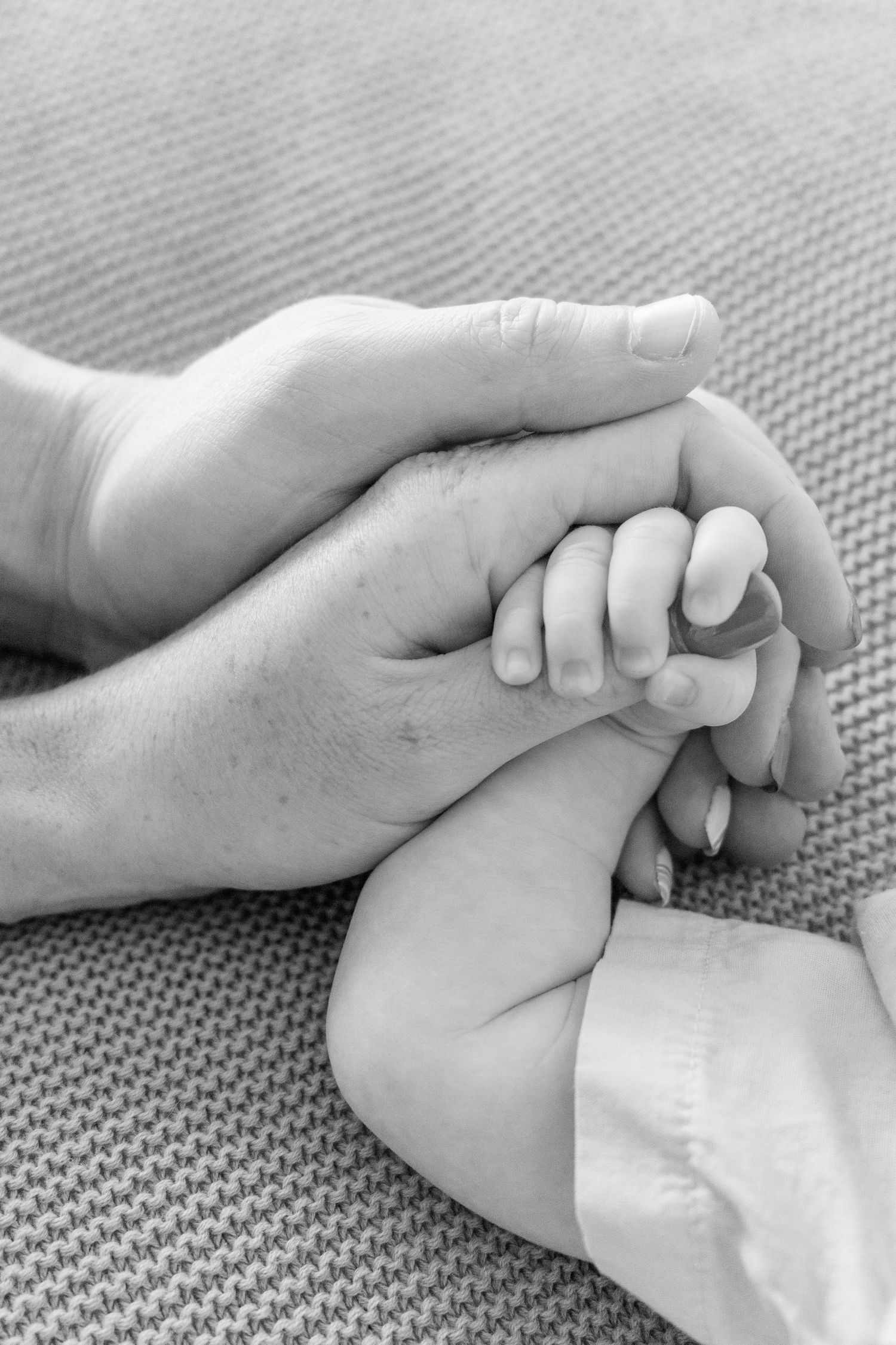Close-up black and white photo of gentle hands holding a tiny infant's hand on textured fabric.
