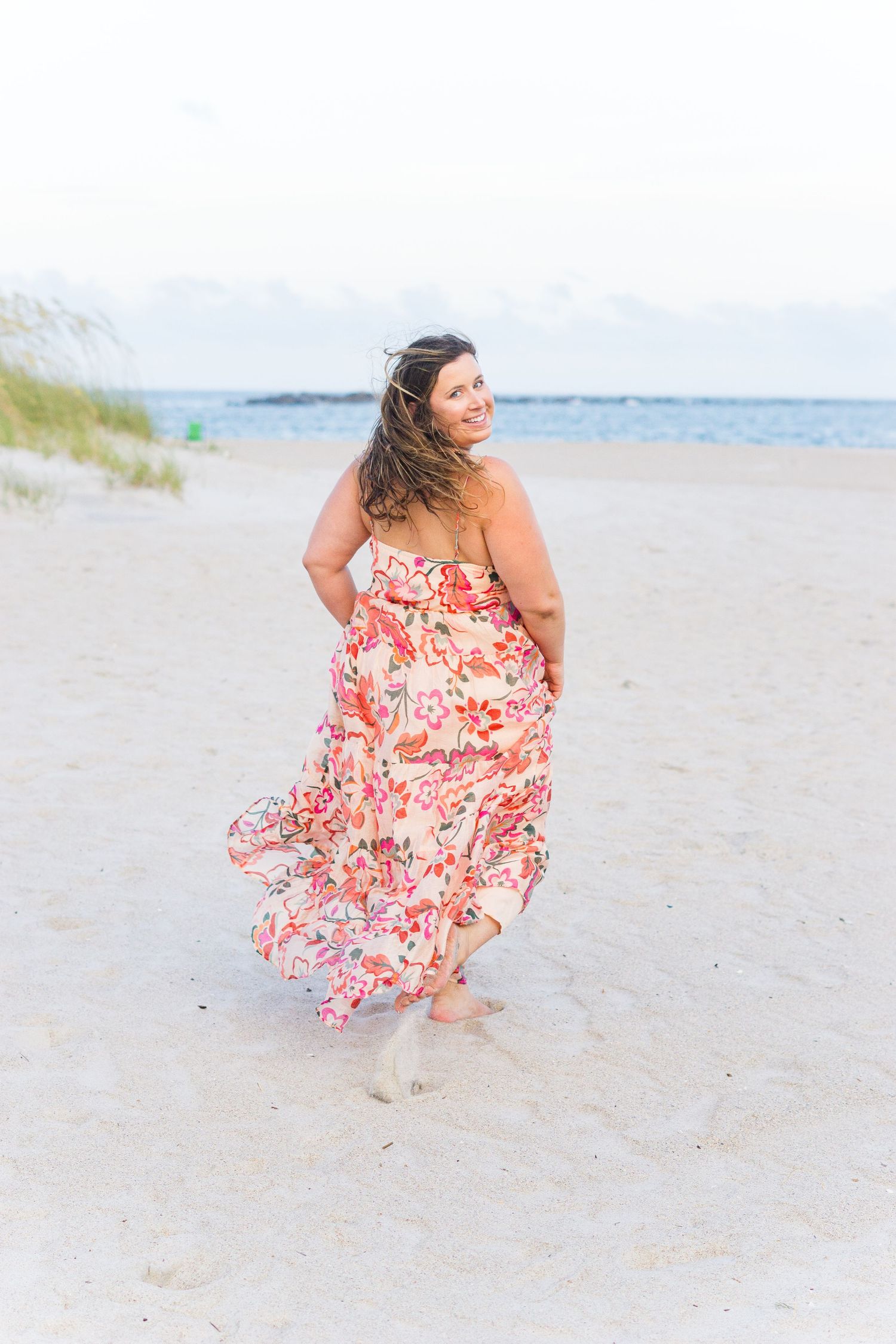 A person in a flowing floral maxi dress twirls on a sandy beach with ocean waves in the background.