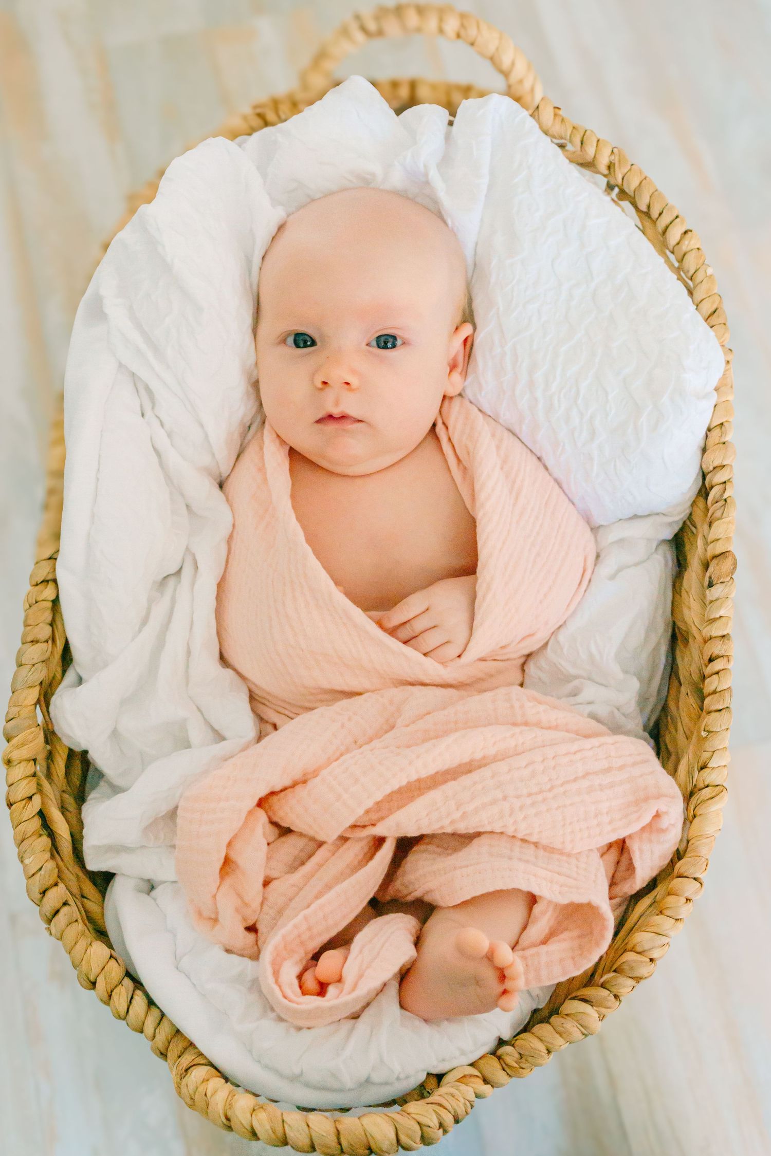 A wide-eyed baby wrapped in peach-colored swaddling lies peacefully in a woven basket with white padding.