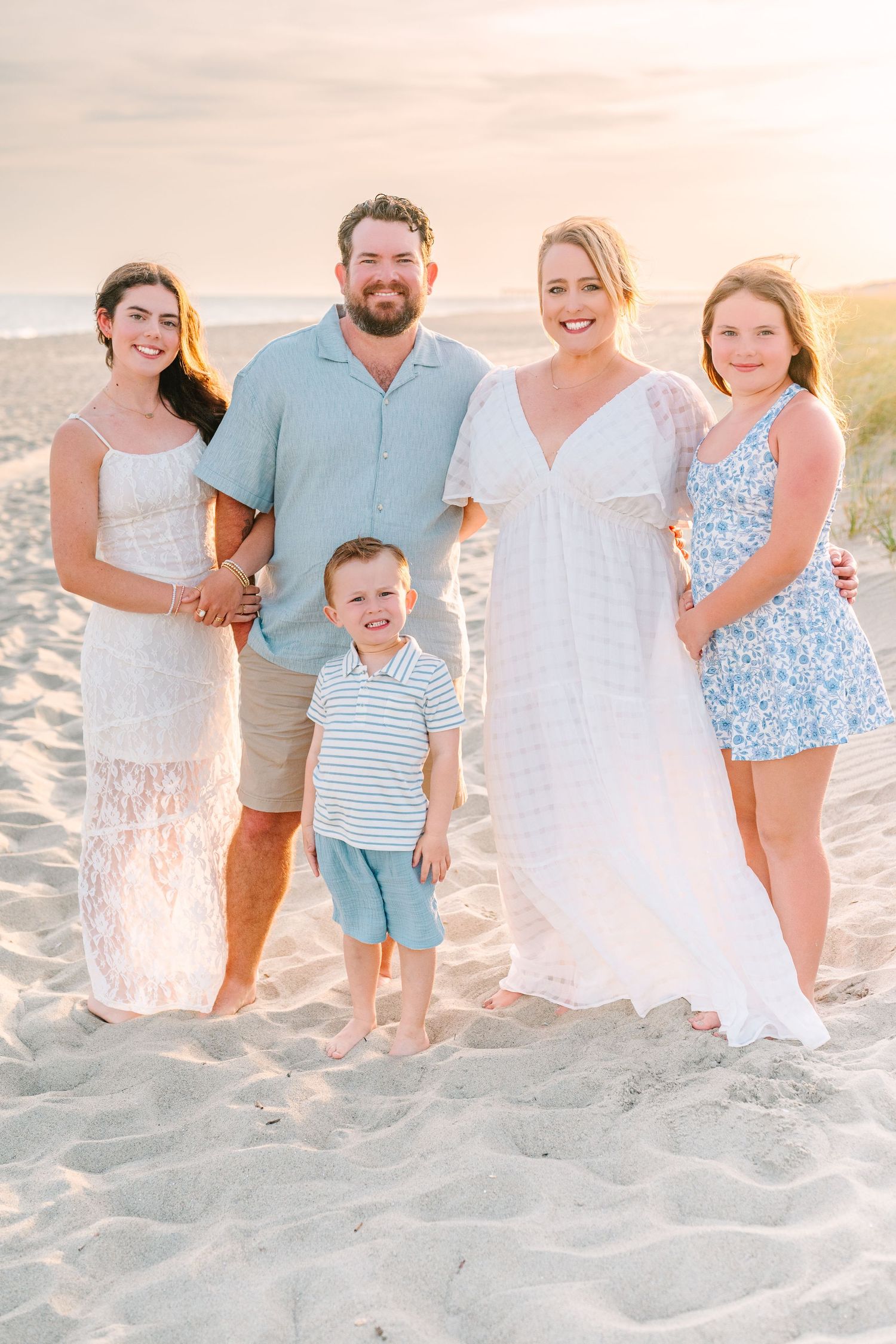 Family group poses together on sandy beach dunes at sunset wearing coordinated beach attire.