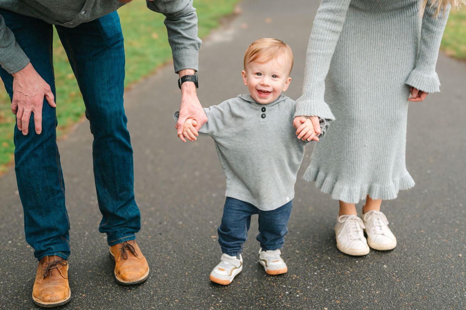 A small child in a gray sweater and jeans holds hands with adults while standing on a paved path outdoors.