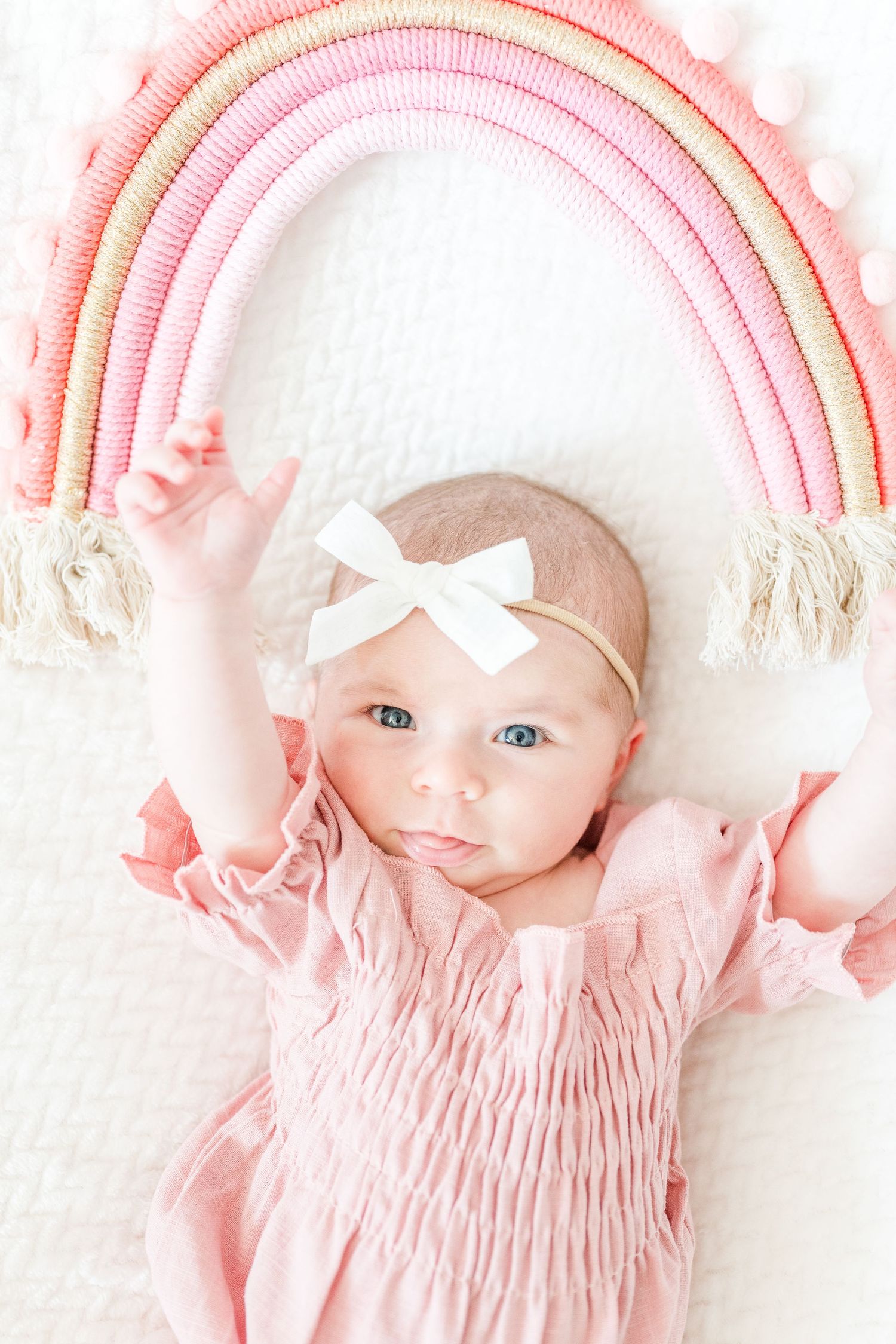 Baby in pink smocked dress and bow headband lying beneath decorative pink macrame rainbow wall hanging.