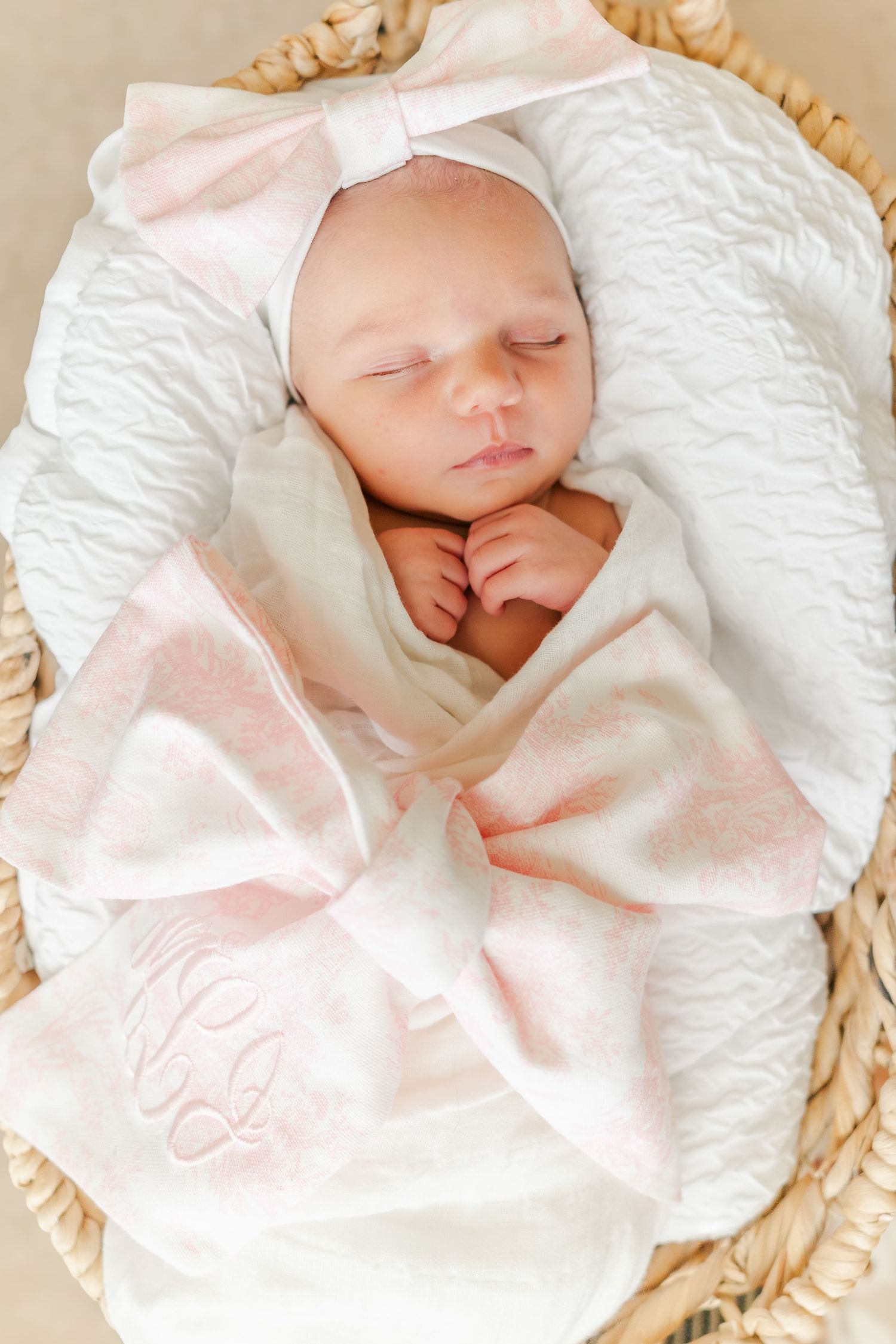 Sleeping newborn baby wearing white with pink bow headband in wicker basket.