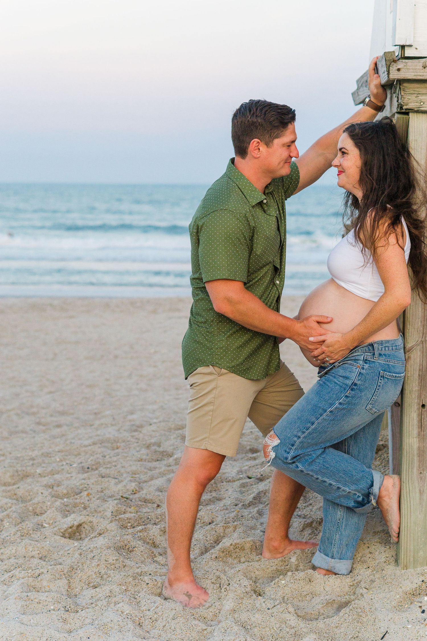 A couple poses playfully against a wooden beach structure during their maternity photoshoot.