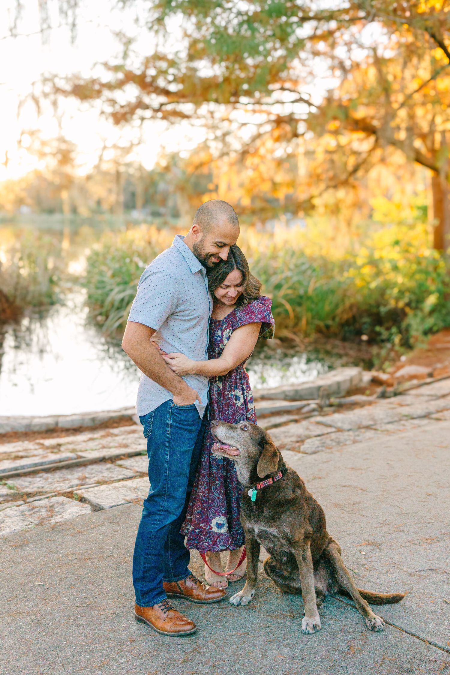 A couple embraces while their loyal dog stands beside them near a scenic waterfront at sunset.