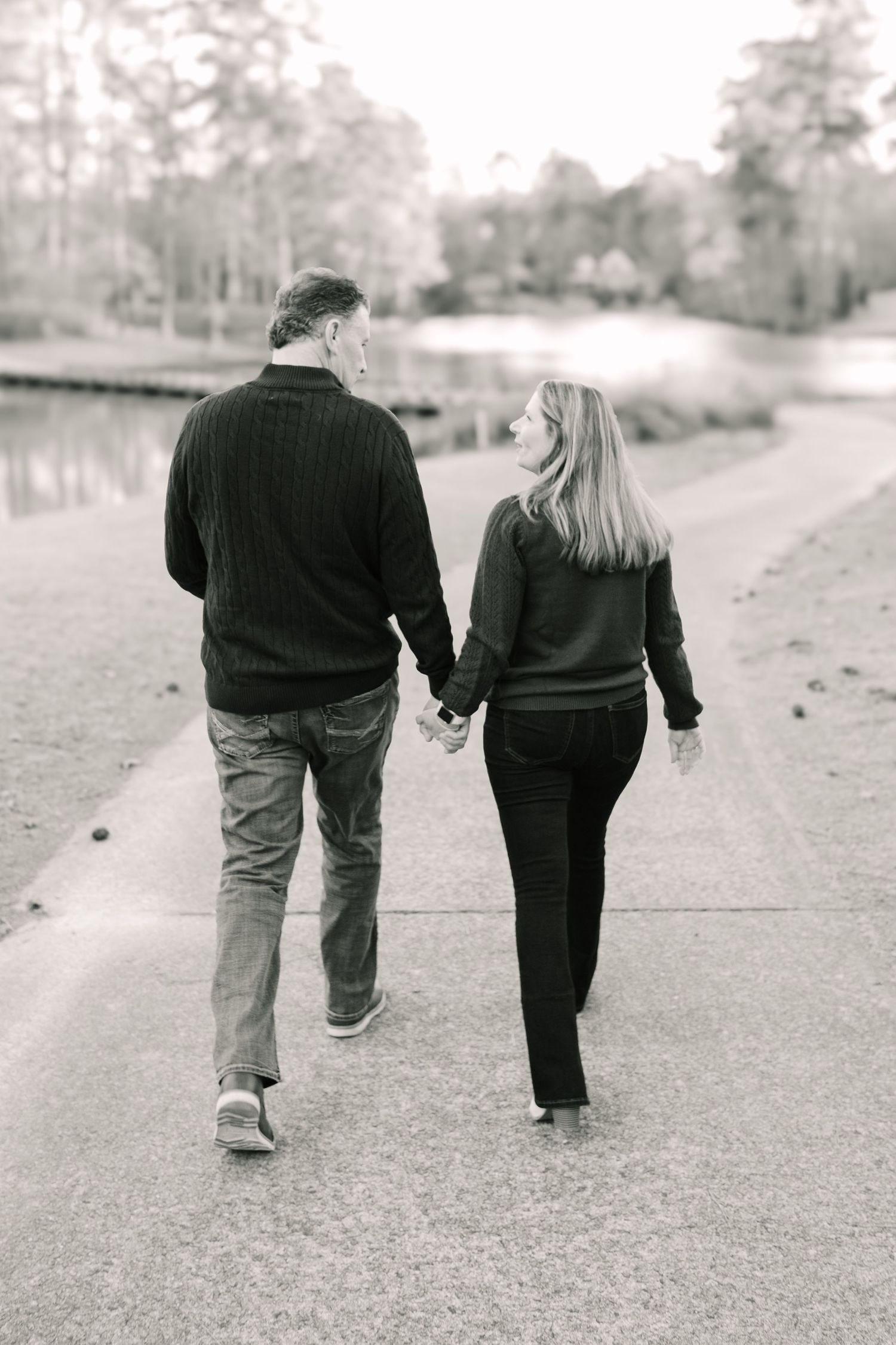A couple walks hand in hand along a peaceful lakeside path in black and white.