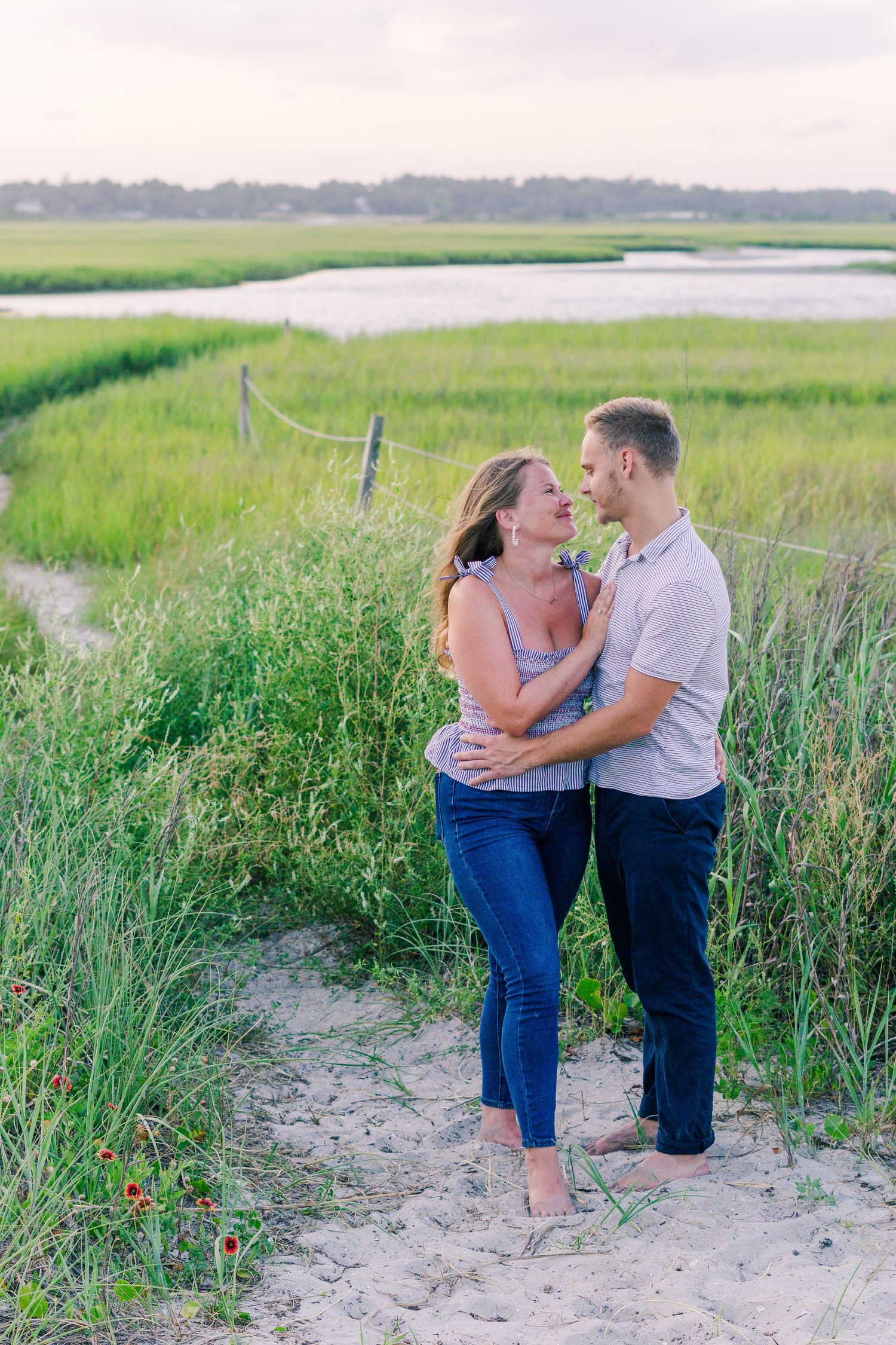 A couple embraces along a coastal marsh path during a romantic photoshoot at sunset.
