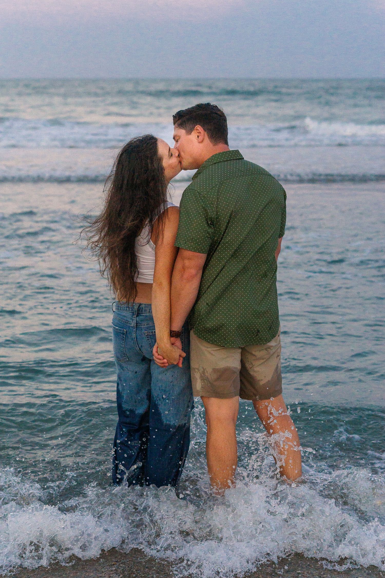 Romantic kiss shared between couple standing in crashing ocean waves at sunset.