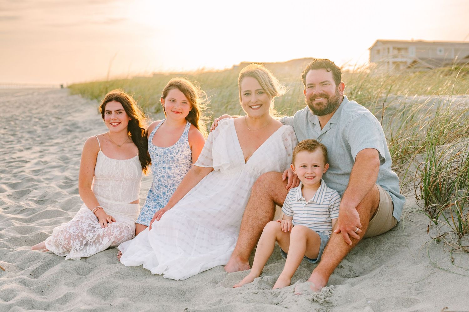 A family sits together on a sandy beach at sunset with coastal dunes in the background.