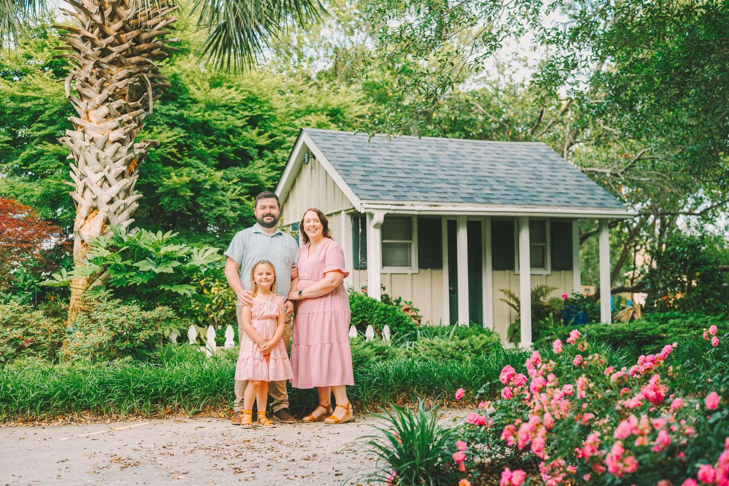 A family stands together in front of a small cottage surrounded by lush gardens and palm trees.