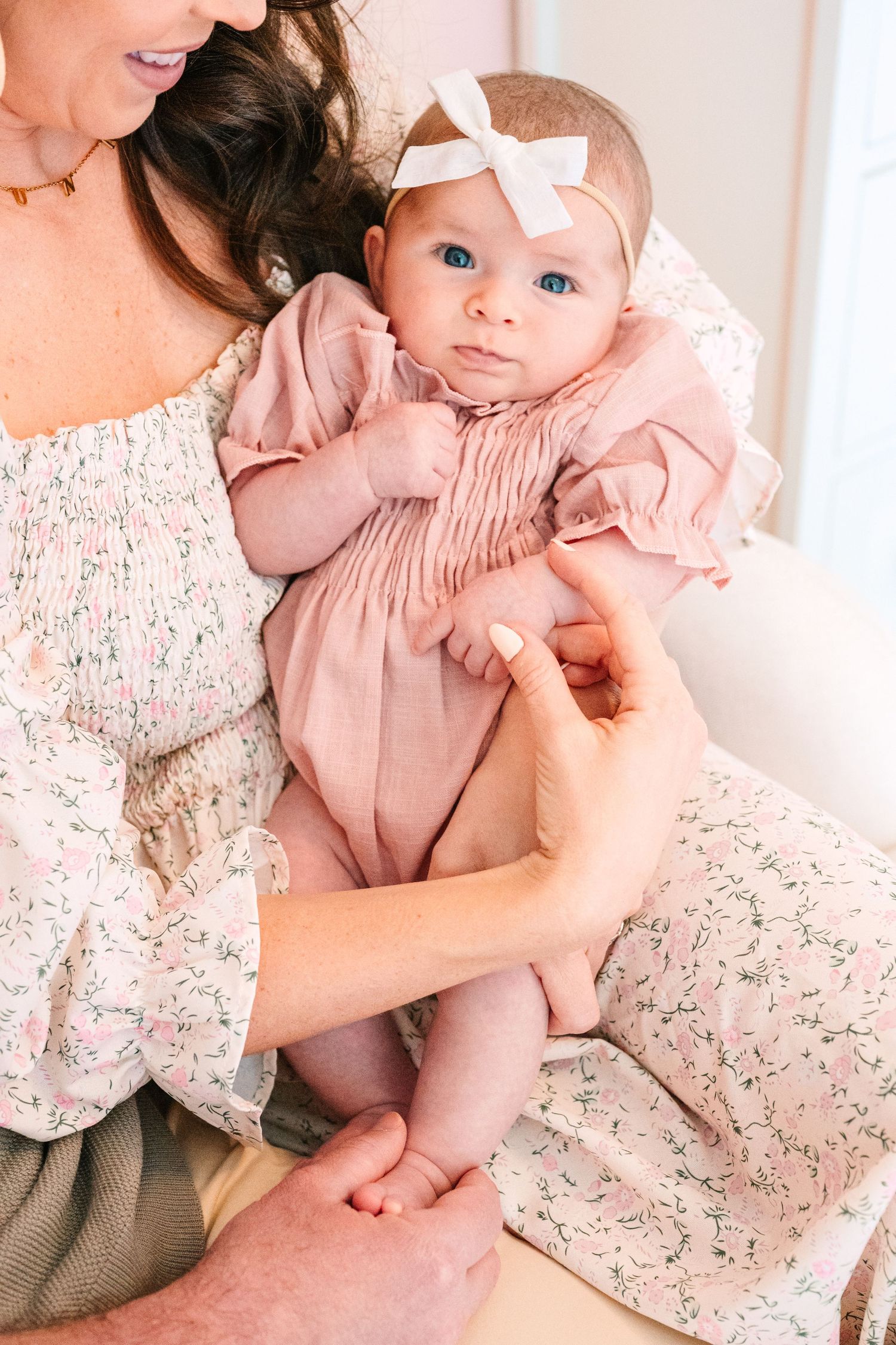 A baby in a pink smocked romper with a white bow headband is held in a floral patterned dress.