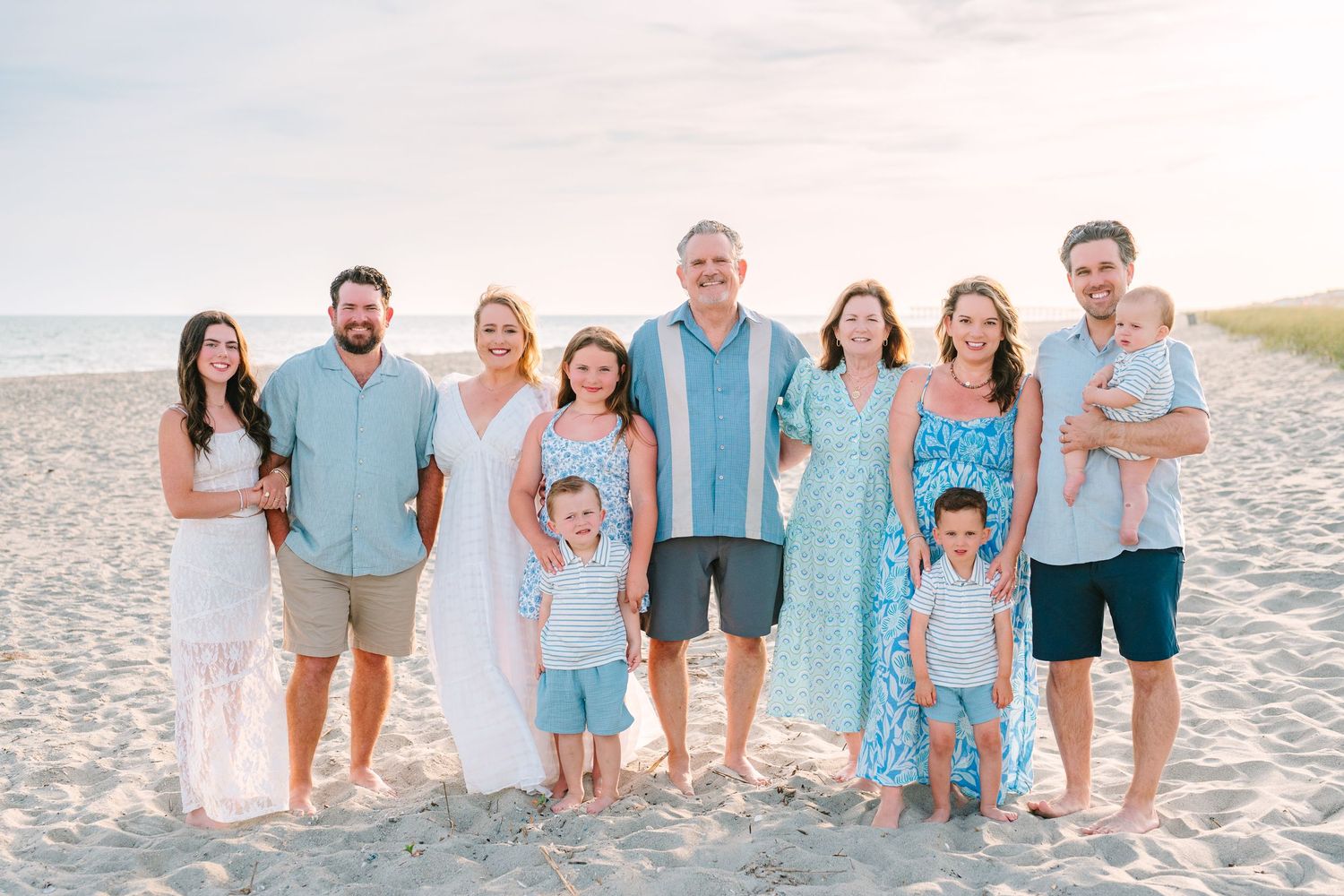 Extended family group wearing coordinated blue and white outfits poses together on a sunny beach at sunset.