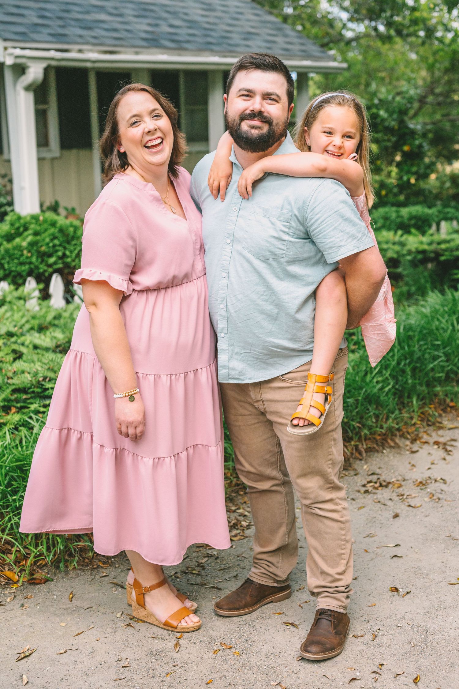 A joyful family of three poses outdoors in coordinated pink and neutral outfits in front of a house.