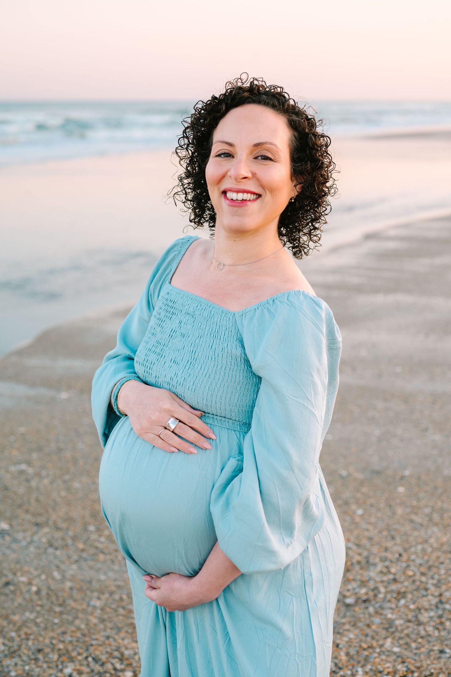 A joyful expectant mother in a flowing turquoise dress stands on a beach at sunset, cradling her baby bump.