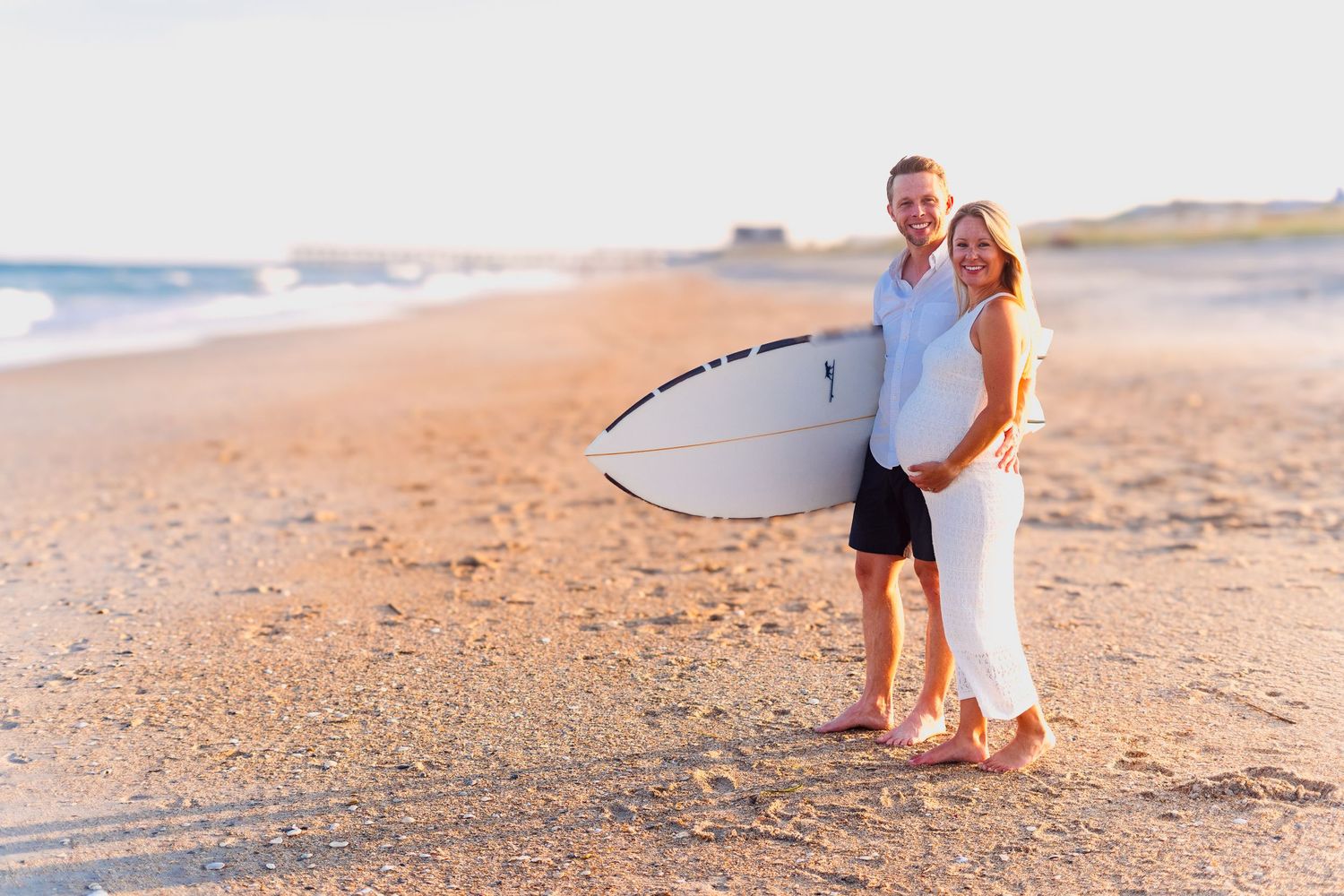Expecting couple poses with surfboard on sandy beach during golden hour.