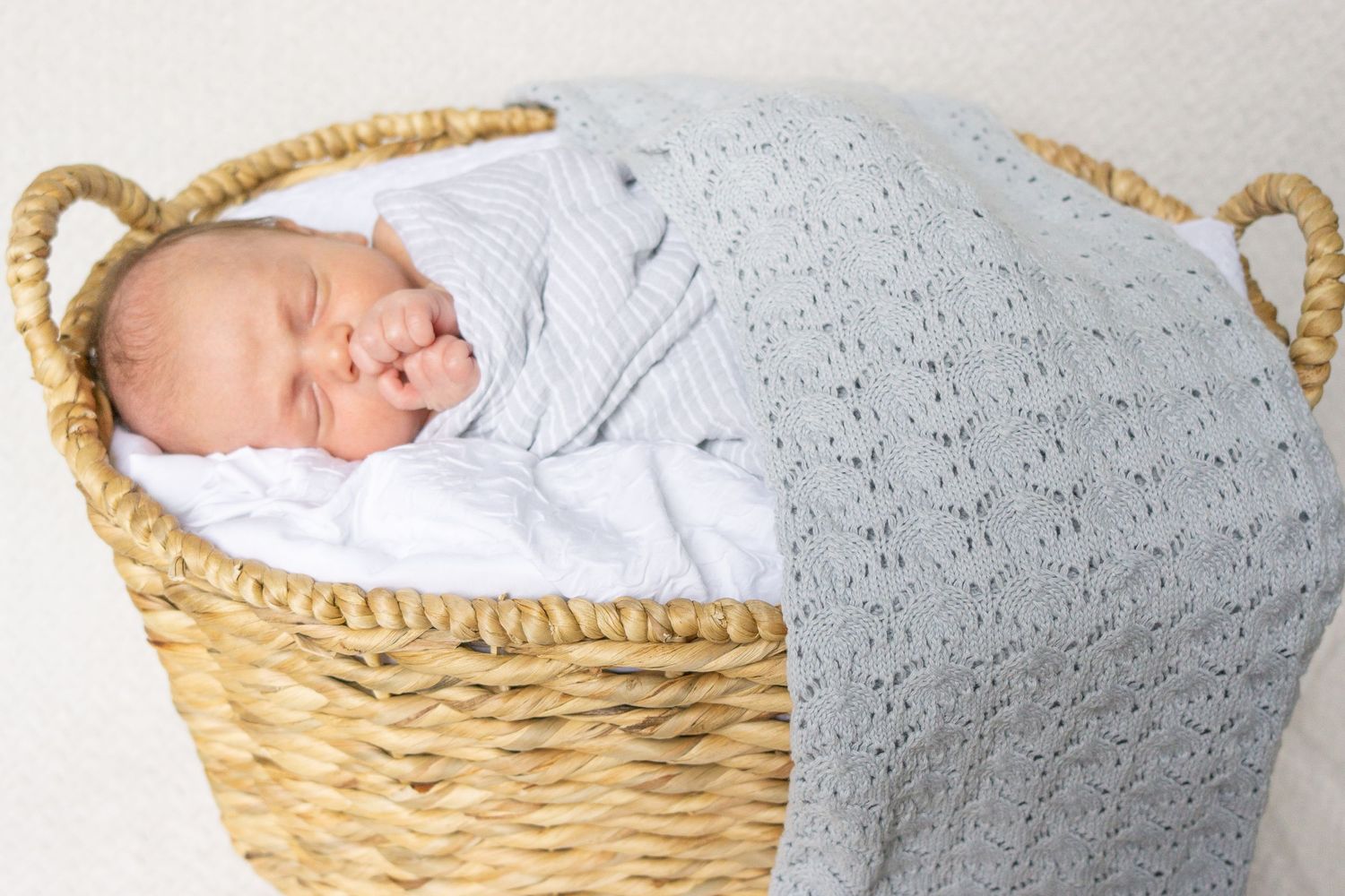 Sleeping newborn in wicker basket covered with gray knit blanket.