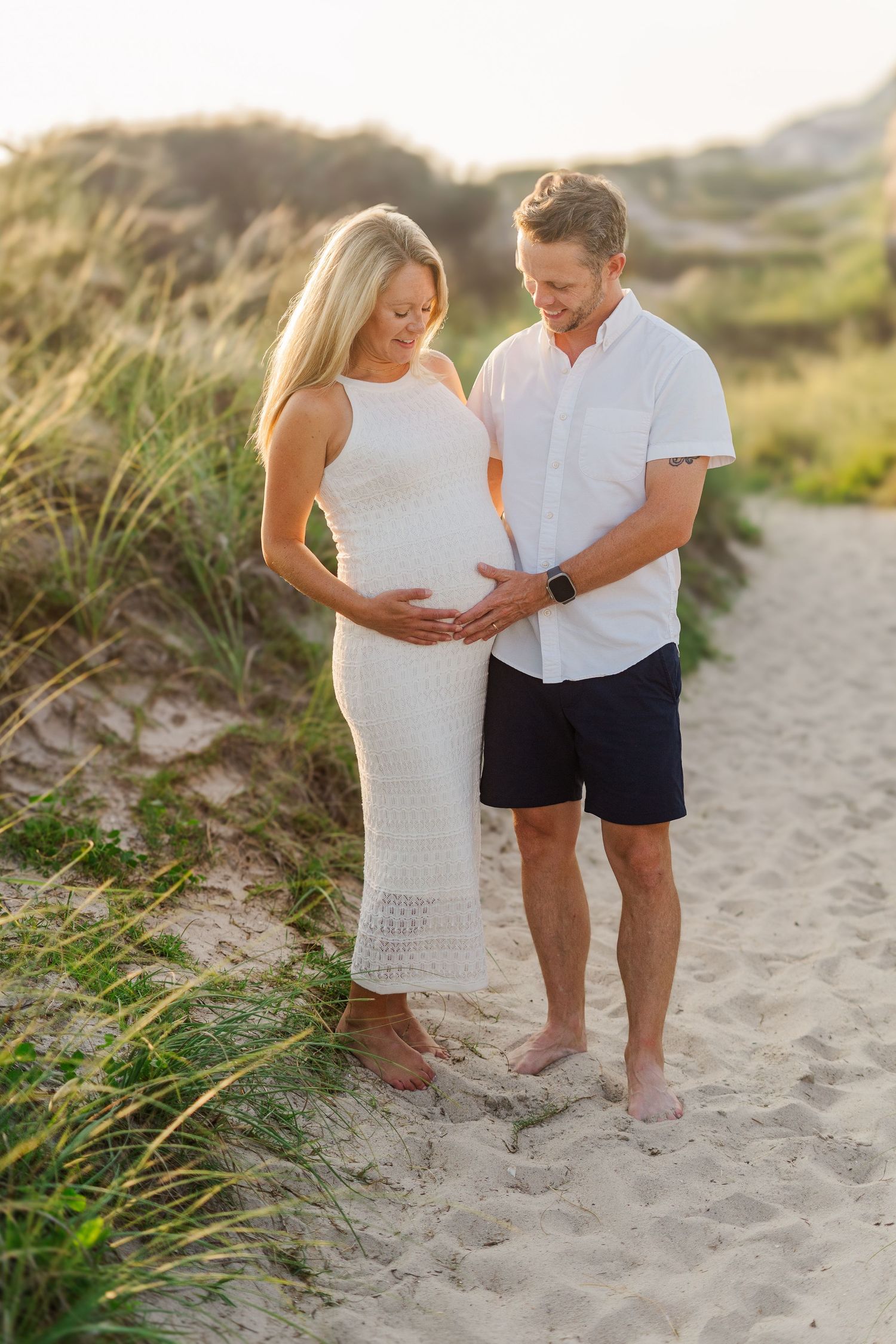 Expectant couple standing together on sandy beach path at sunset.