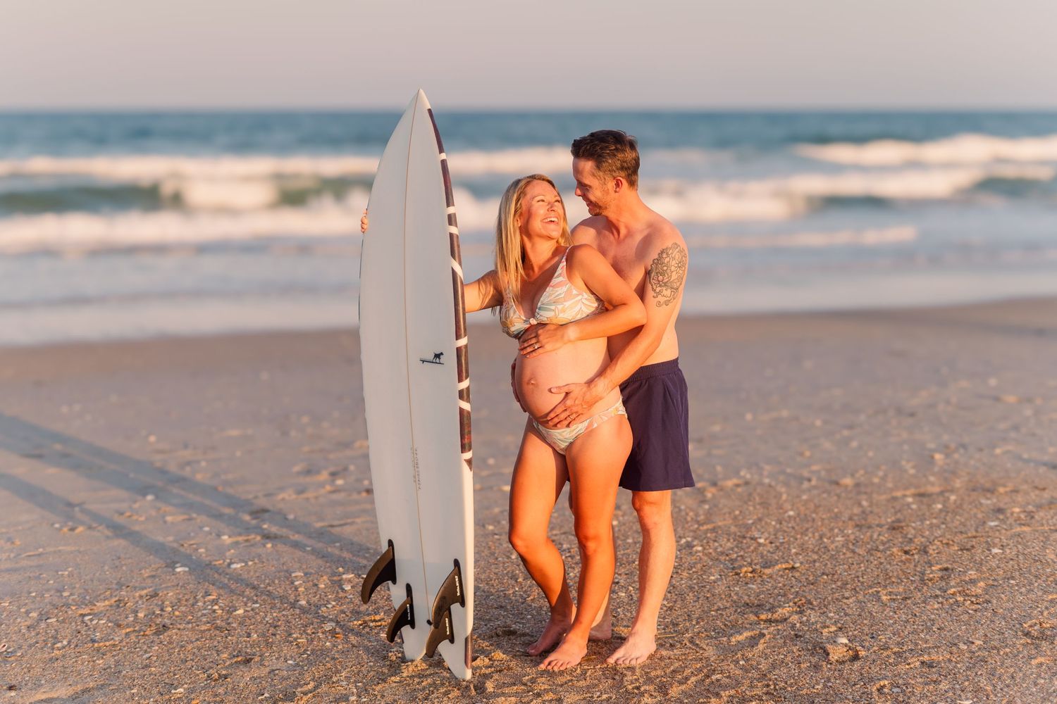 Couple poses together on beach with white surfboard during sunset.