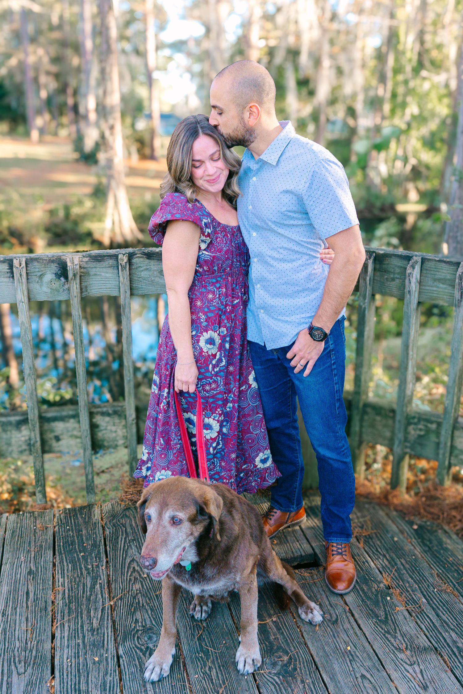 Couple with their dog posing on wooden deck overlooking water and cypress trees.