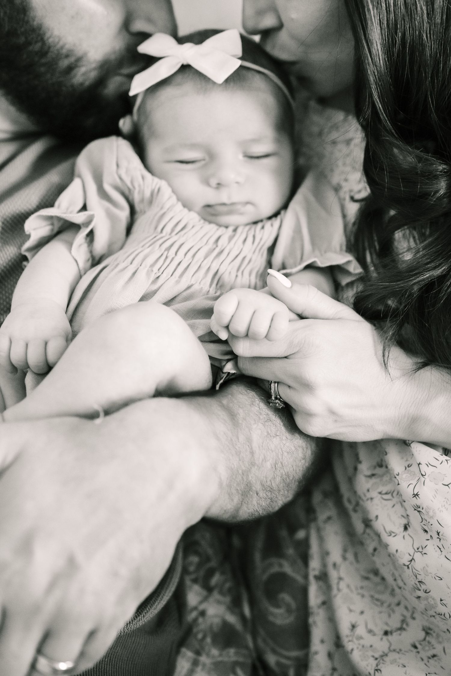 A black and white portrait of parents lovingly cradling their sleeping infant in a white bow headband.